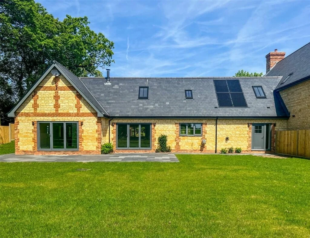 Back of a brick house with a dark gray roof, solar panels, and several skylights, overlooking a green lawn and a patio area.