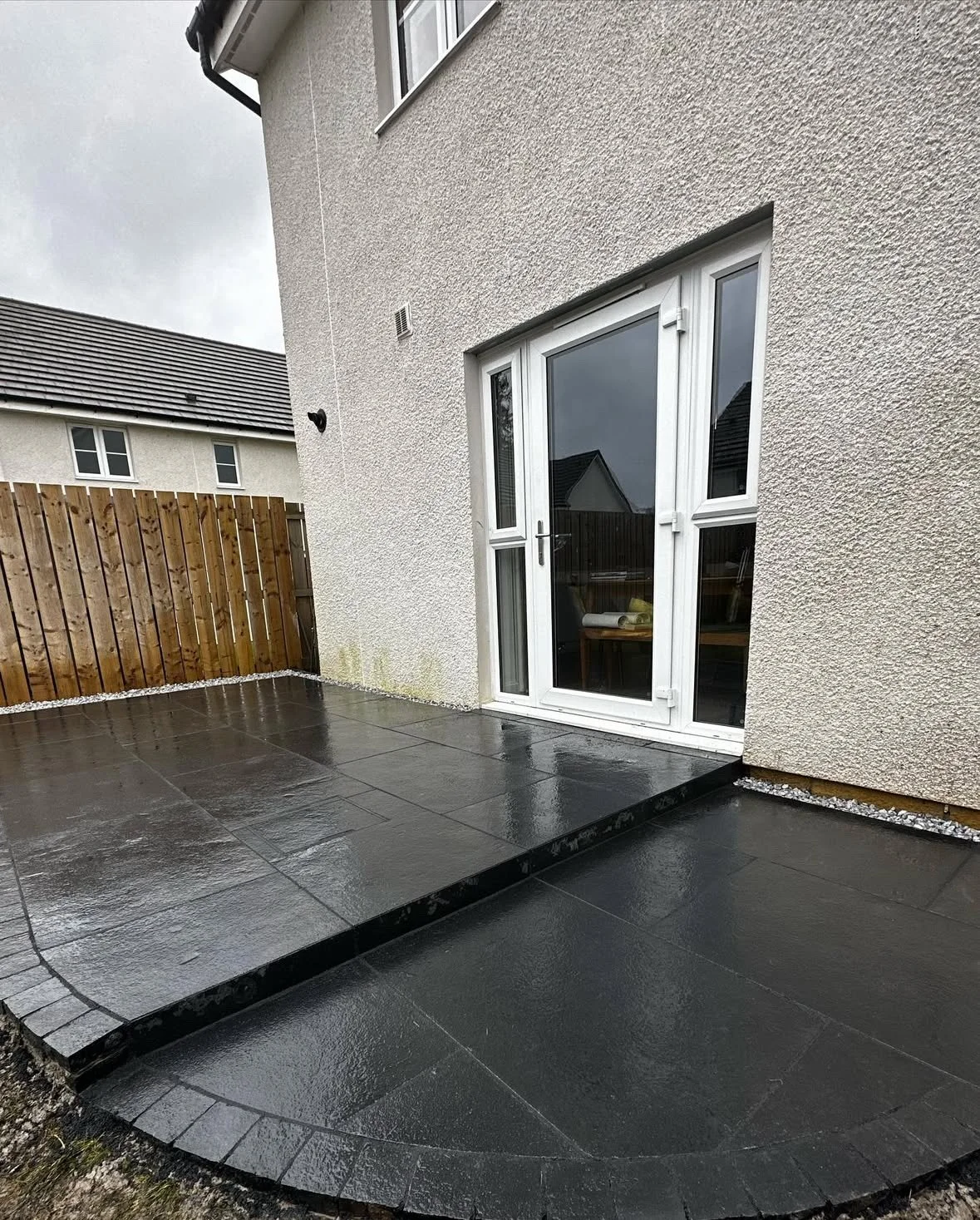 Wet black patio tiles outside of a house with a white sliding glass door and a wooden fence in the background.