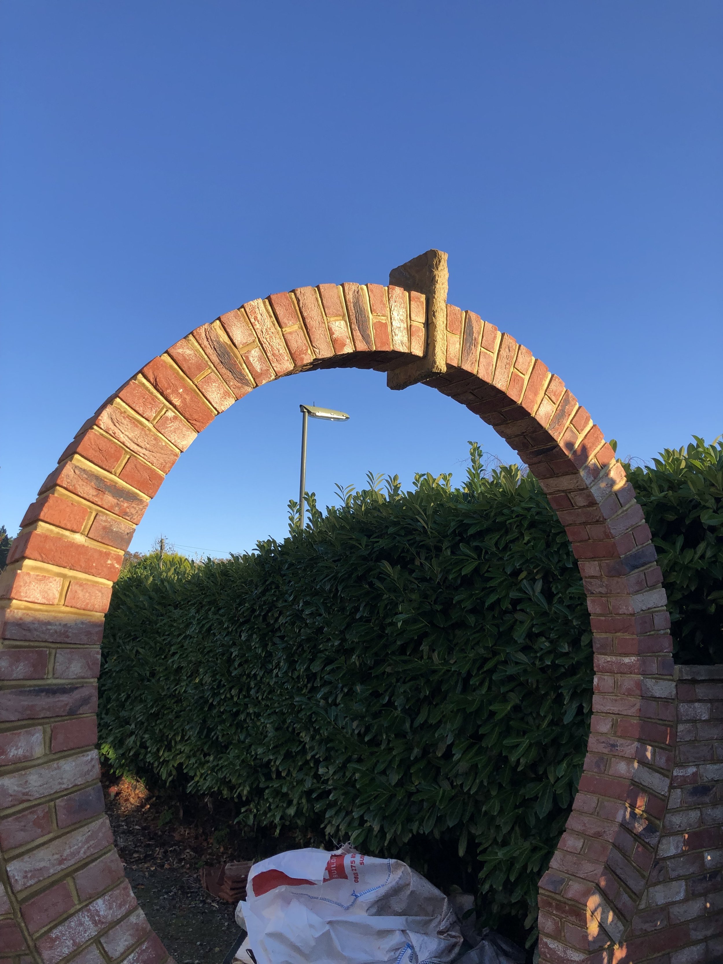 Brick archway with a section missing, set against a green hedge and a clear blue sky, with a streetlamp visible behind.