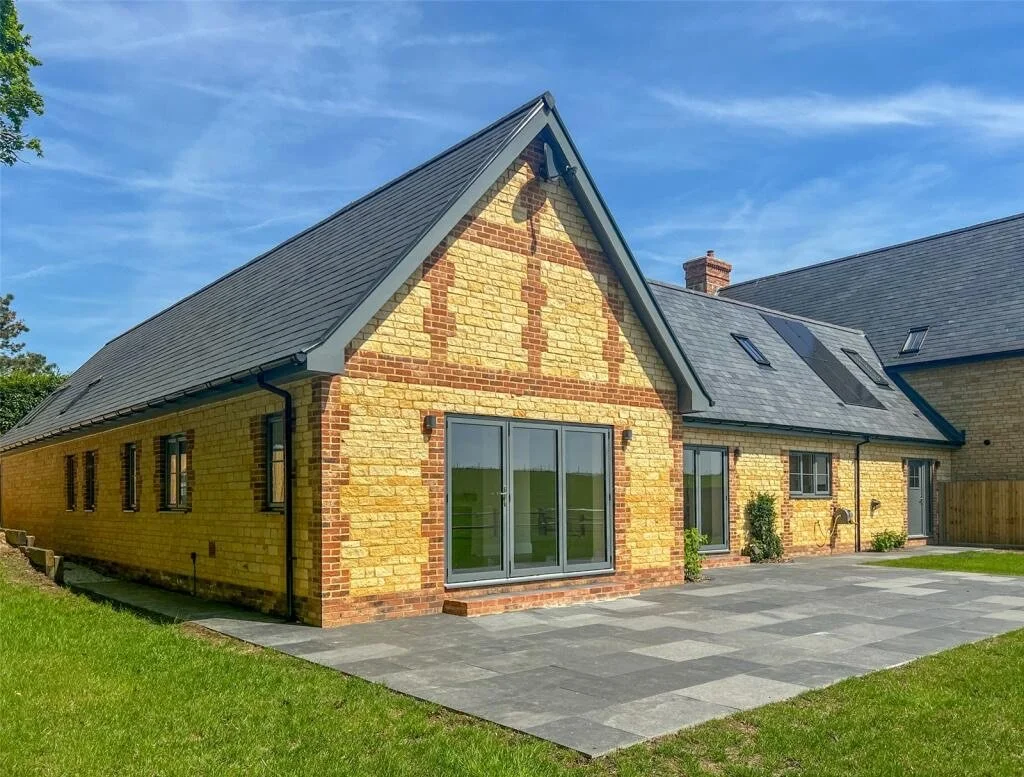 Backyard view of a yellow brick house with a gray shingled roof and sliding glass doors, surrounded by a patio and green lawn, under a blue sky.