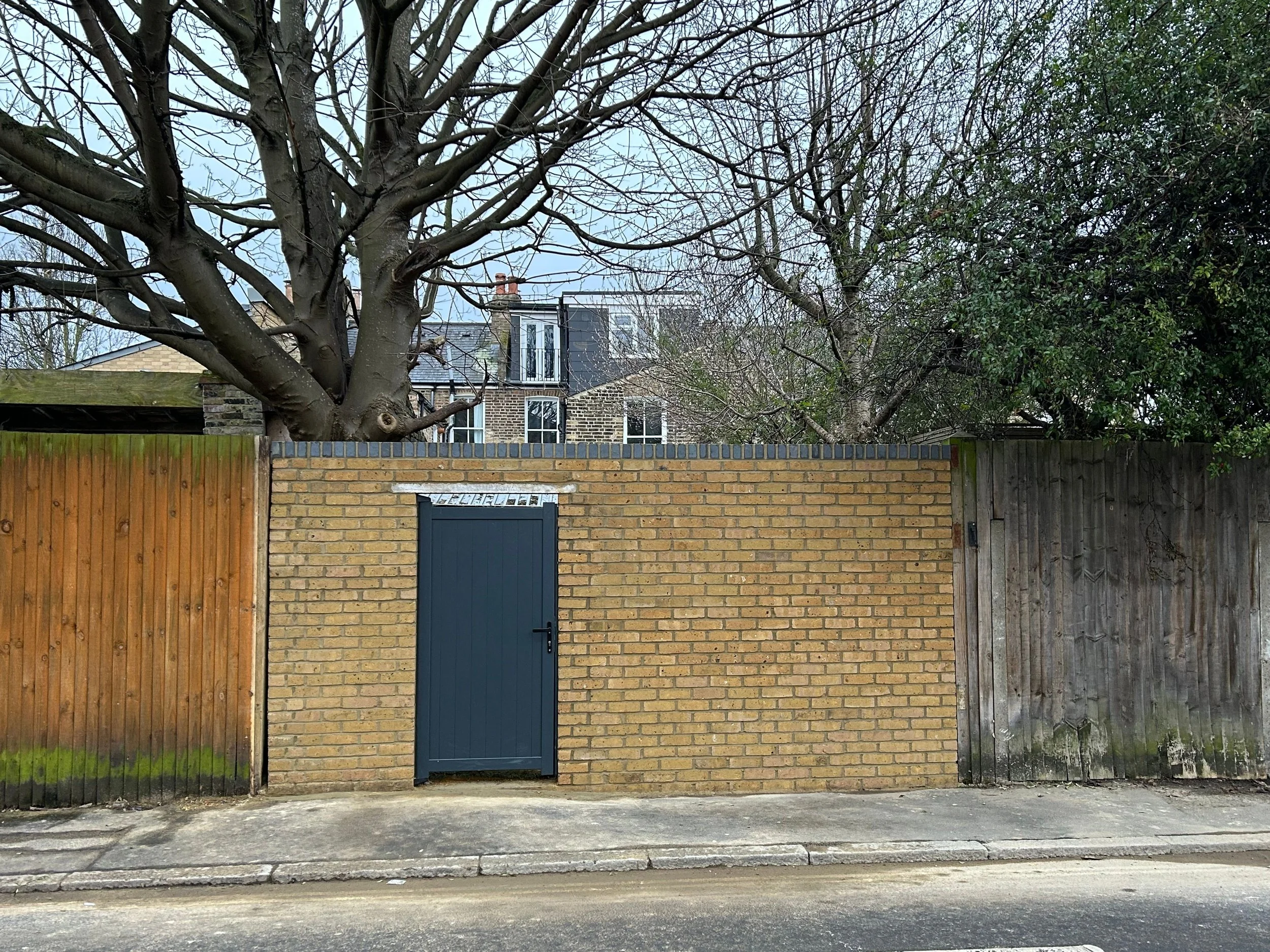 A brick wall with a blue door, flanked by wooden and brick fencing, above which are leafless trees and a multistory brick building in the background.