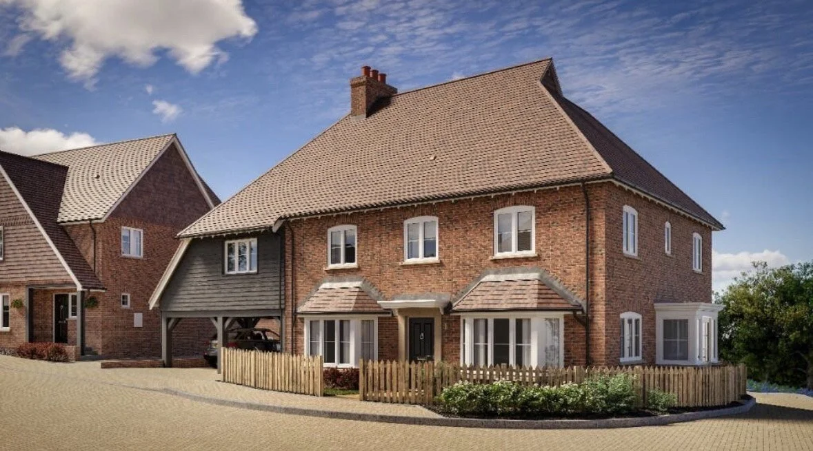 A large brick house with a steep roof and multiple windows, surrounded by a small garden and wooden fence, under a partly cloudy sky.