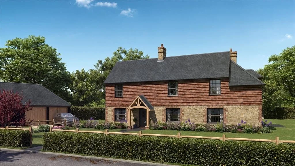 A historic two-story brick house with a grey shingle roof and a small front porch, surrounded by a well-maintained garden and a wooden fence, with a smaller outbuilding and trees in the background under a blue sky.