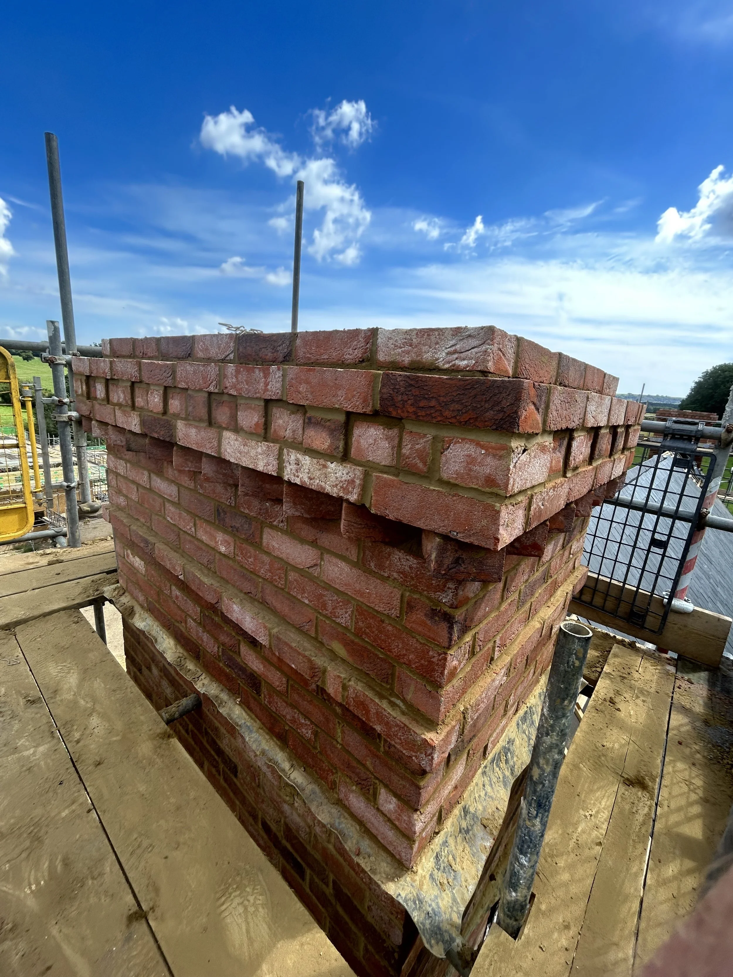 Close-up of a brick chimney under construction on a building roof with scaffolding, in a partly cloudy sky.