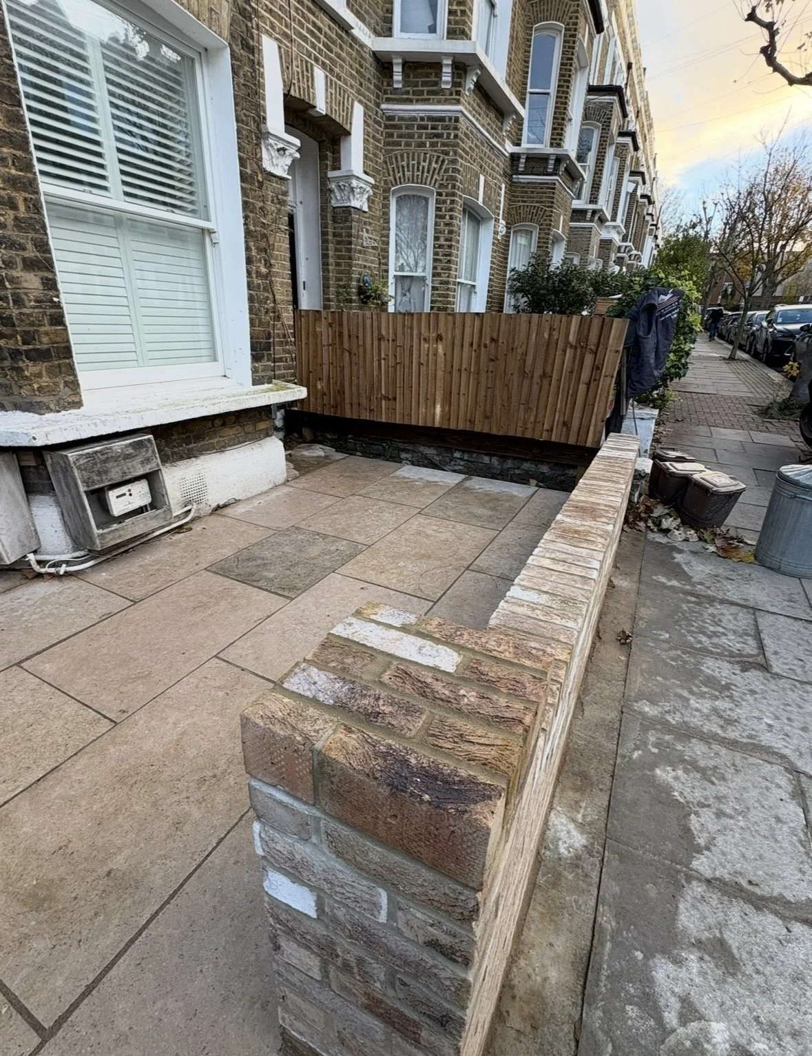 Small outdoor patio with brick wall and wooden fence in front of brownstone building, featuring chairs and parked cars on the street.