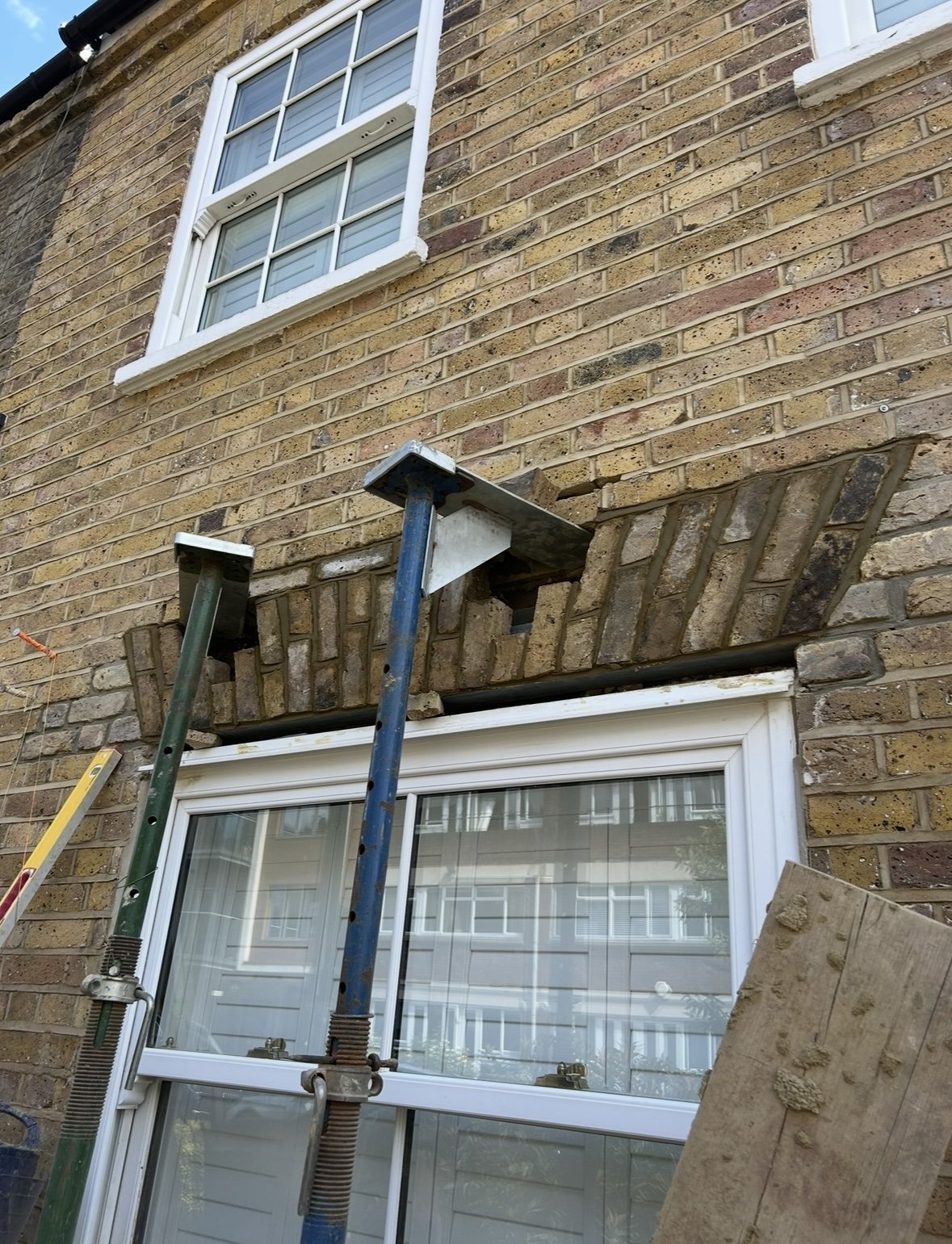 A brick house with a white-framed window on the upper floor and a lower window being replaced or repaired with scaffolding in front.