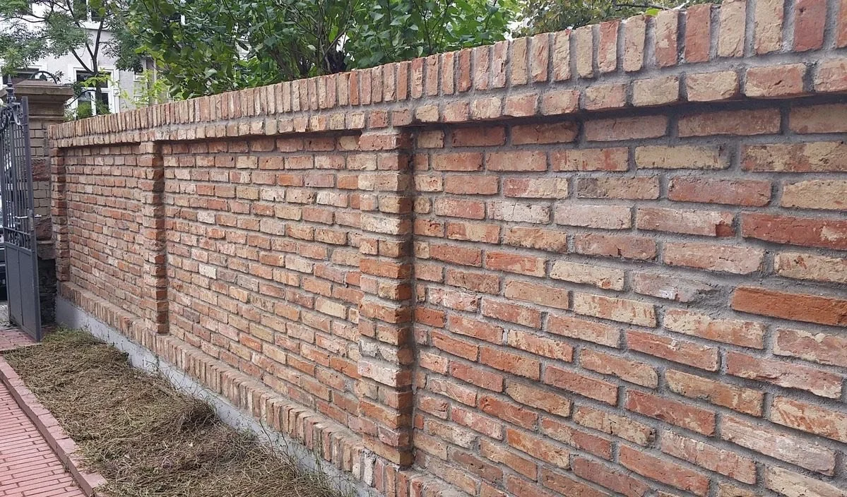 A long brick wall with three rectangular indentations, surrounded by a sidewalk with some dirt and grass along the base.