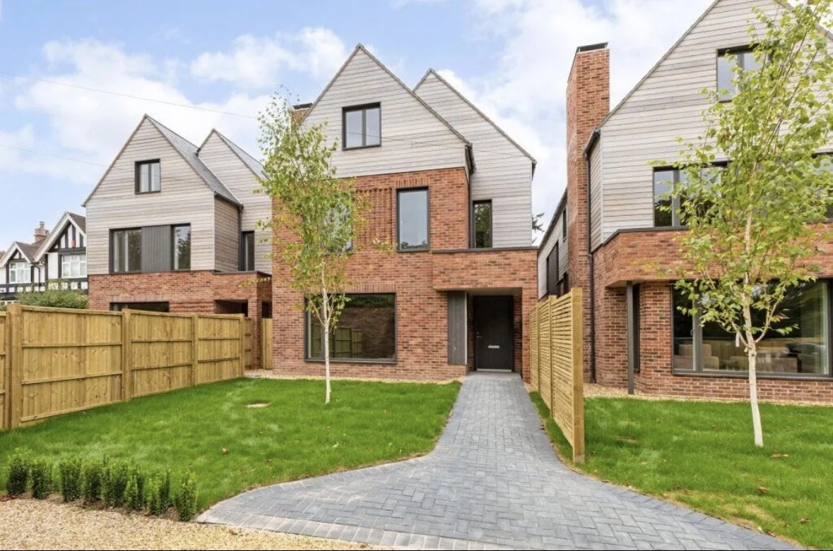 Modern townhouse with brick and gray siding, small front yard with green grass, young trees, paved walkway, and wooden fences.