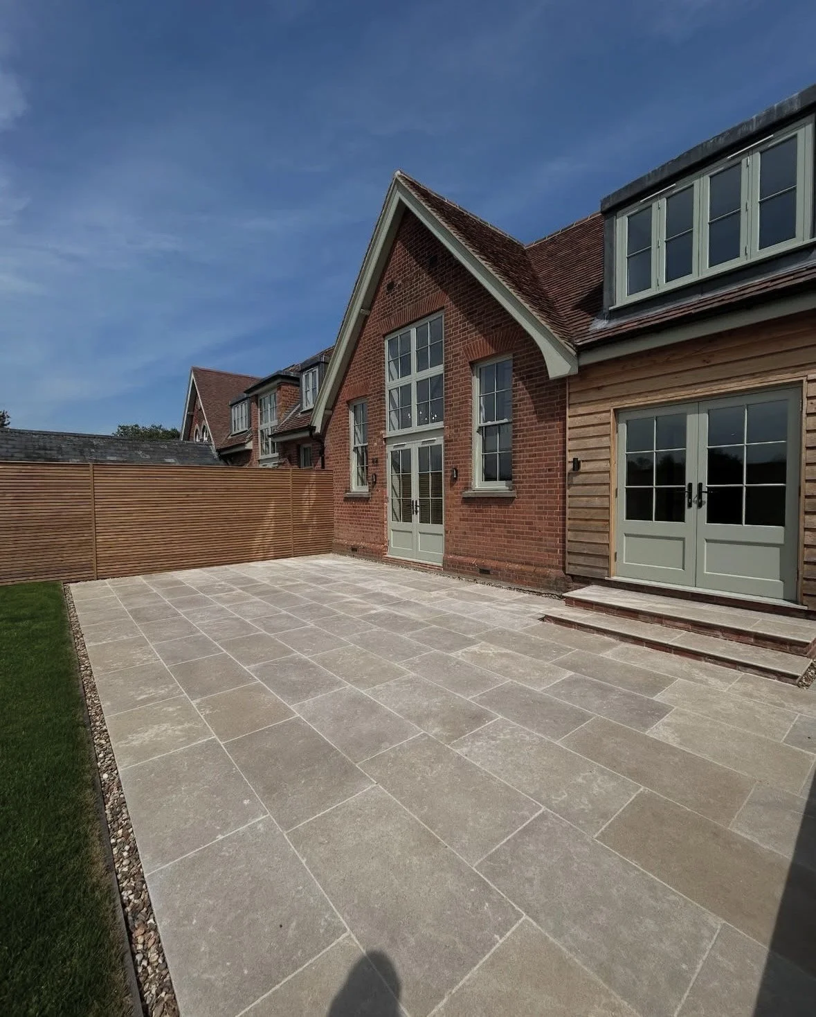Backyard patio with large beige stone tiles, a wooden privacy fence on the left, a red brick house with green window and door frames, and a grassy lawn on the left side under a blue sky.