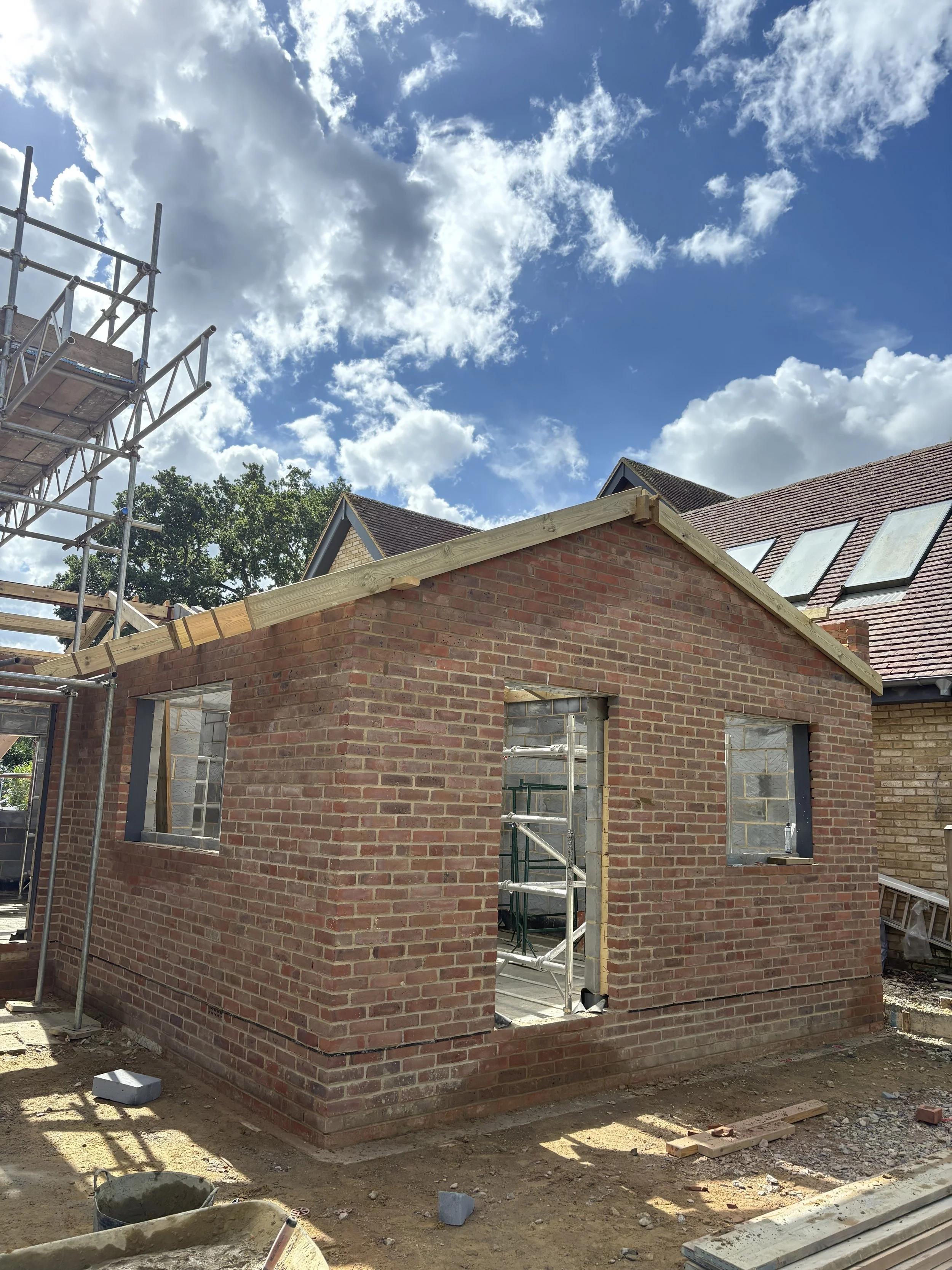 A small brick building under construction with scaffolding nearby, open doorway, and window openings, set against a partly cloudy sky.
