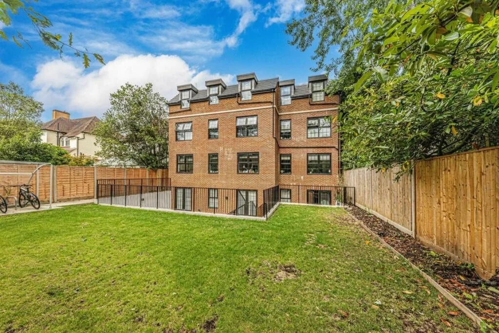 Backyard with green grass, wooden fence, and exterior of a brick apartment building with multiple windows and a decorative roof.