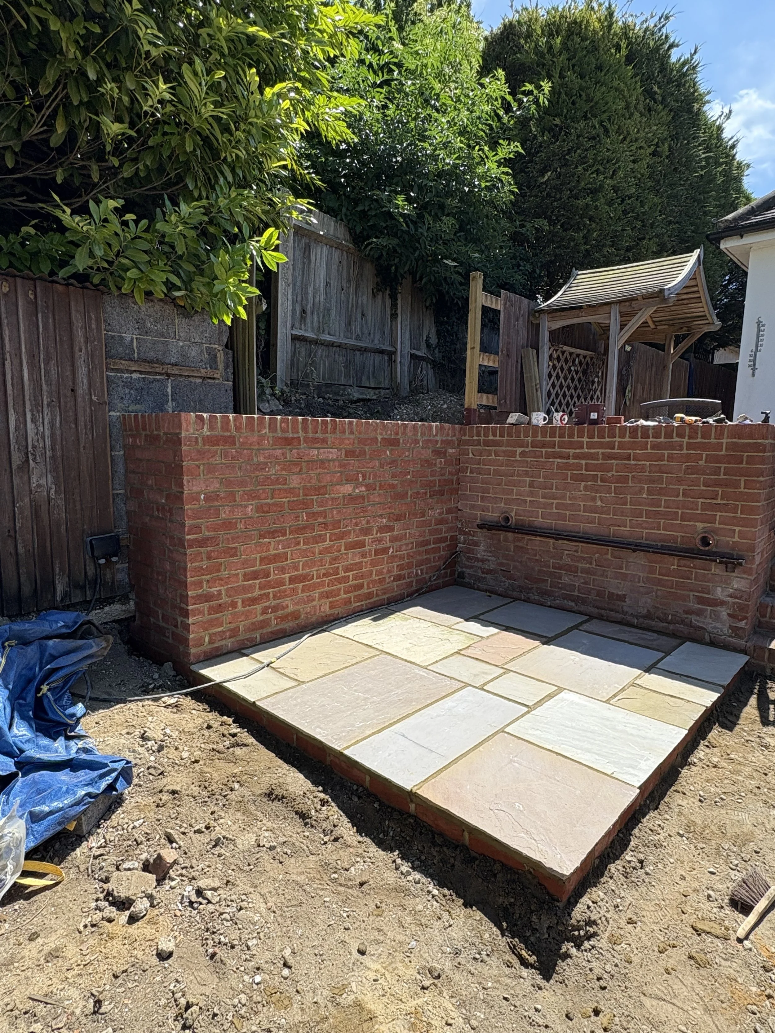 Construction site with a partially built brick wall and a paved area with large, multi-colored stone tiles, surrounded by a wooden fence, trees, and a small wooden roof structure.
