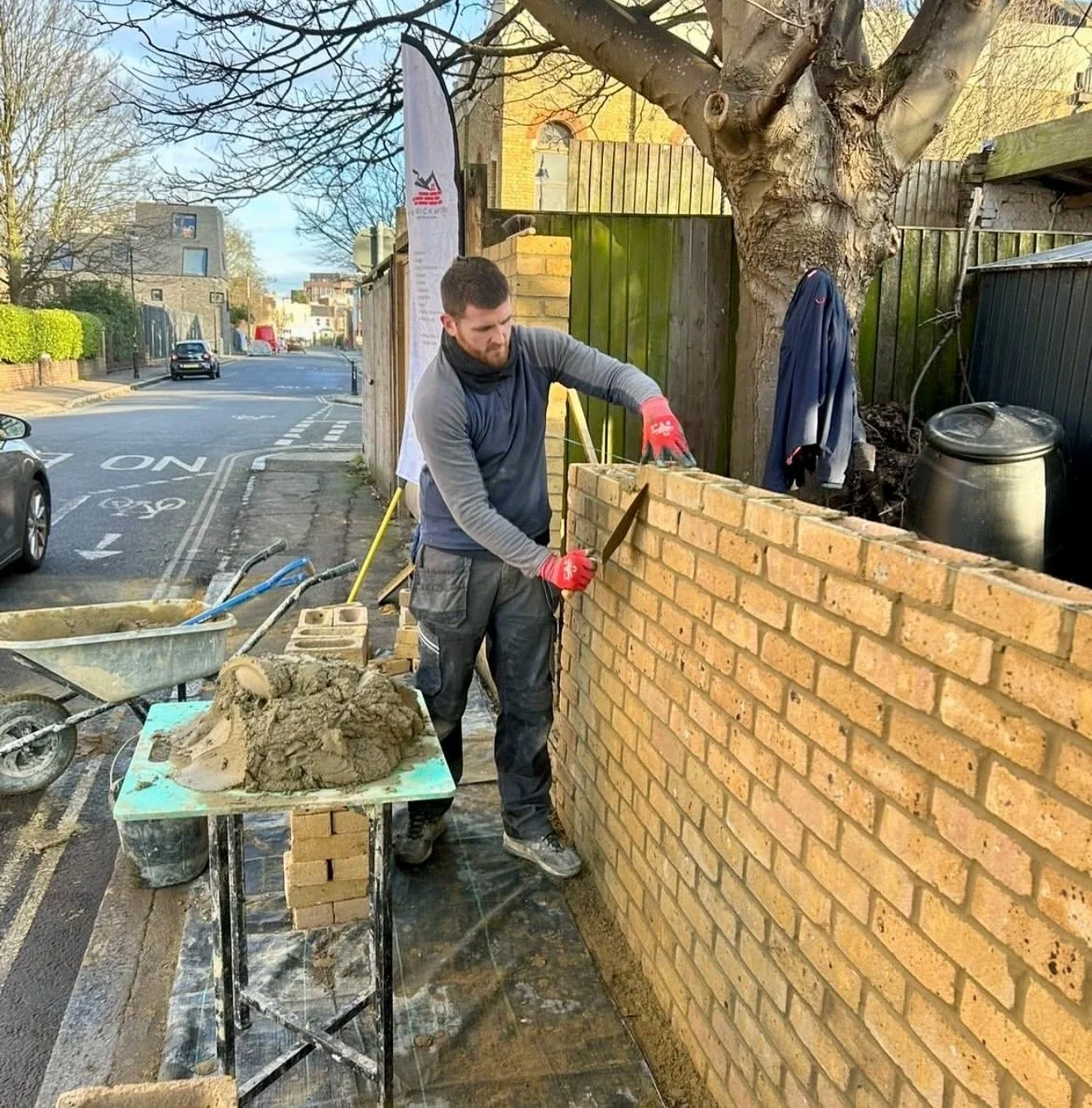 A man working outdoors on a brick wall construction, wearing red gloves, black pants, a long sleeve shirt, and a vest. He is holding a trowel and working on the brick wall. There are construction tools and material, including a wheelbarrow with cement, nearby. The scene takes place on a street with cars and houses in the background, under a partly cloudy sky.