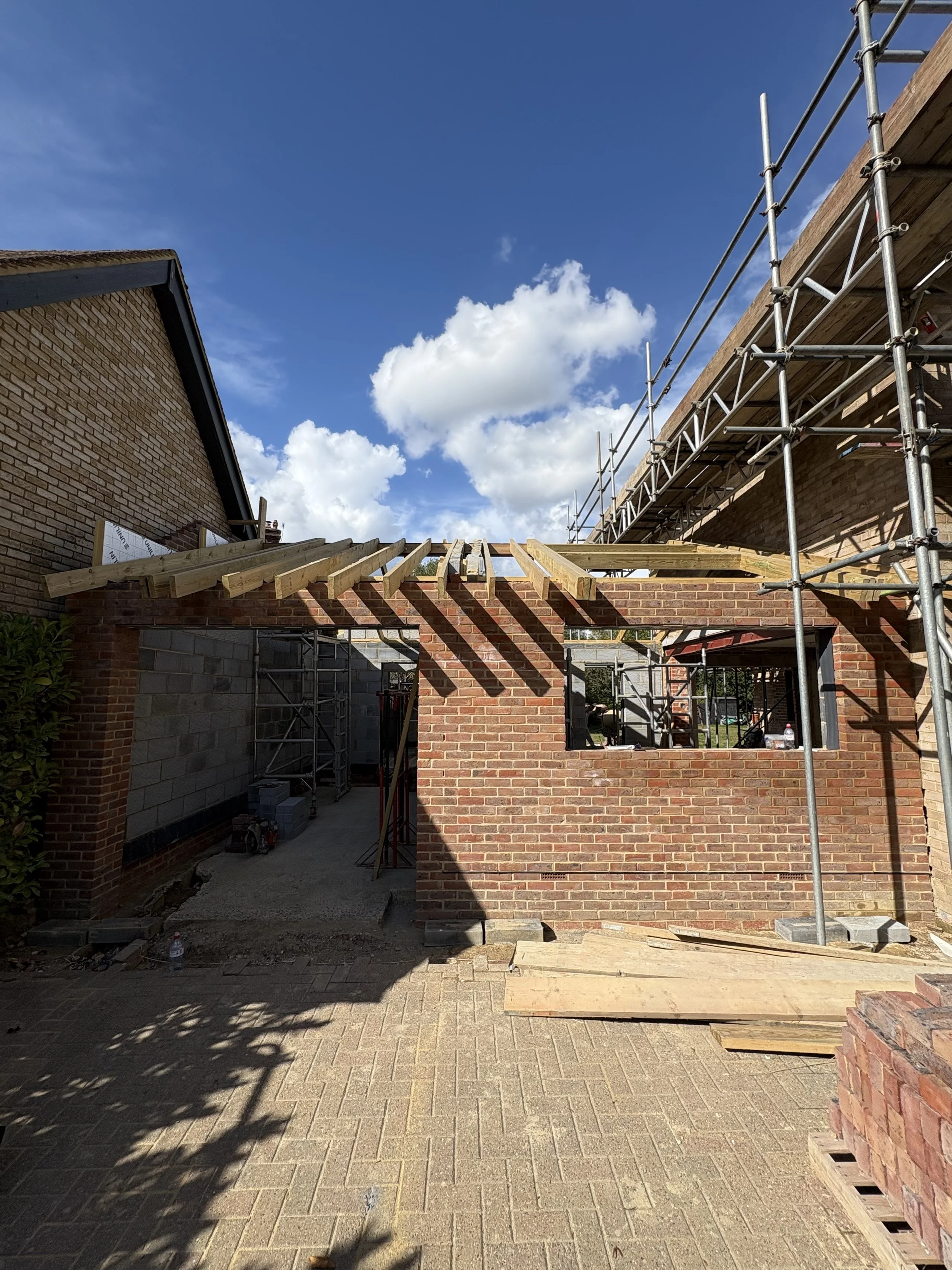Construction site showing brick walls, scaffolding, and wooden beams being installed for building extension, with a clear blue sky and some clouds in the background.