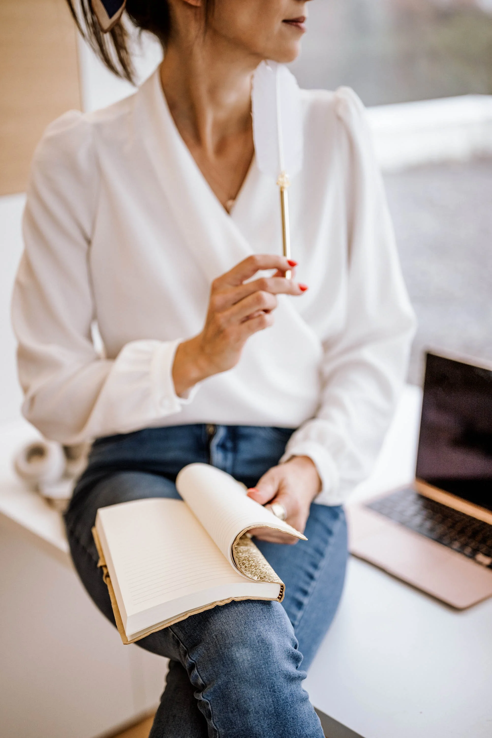 Eine Frau sitzt mit einem offenen Notizbuch auf dem Schoß, hält einen Stift in der Hand und schaut nach rechts. Neben ihr liegt ein Laptop.