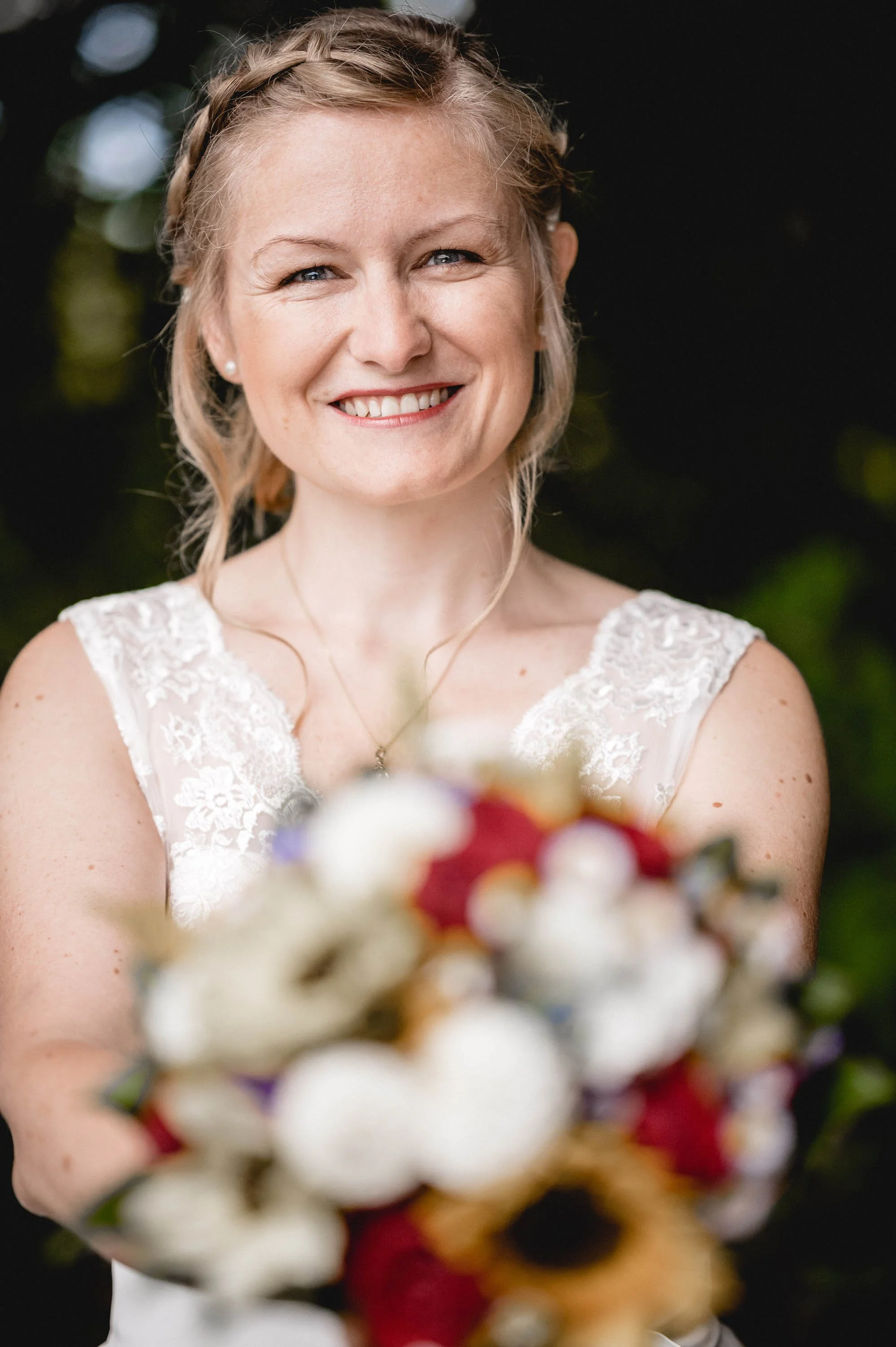 Eine lachende Frau im weißen Brautkleid hält einen Blumenstrauß. Sie hat blonden, leicht gewellten Haaren und trägt kleine Ohrringe.