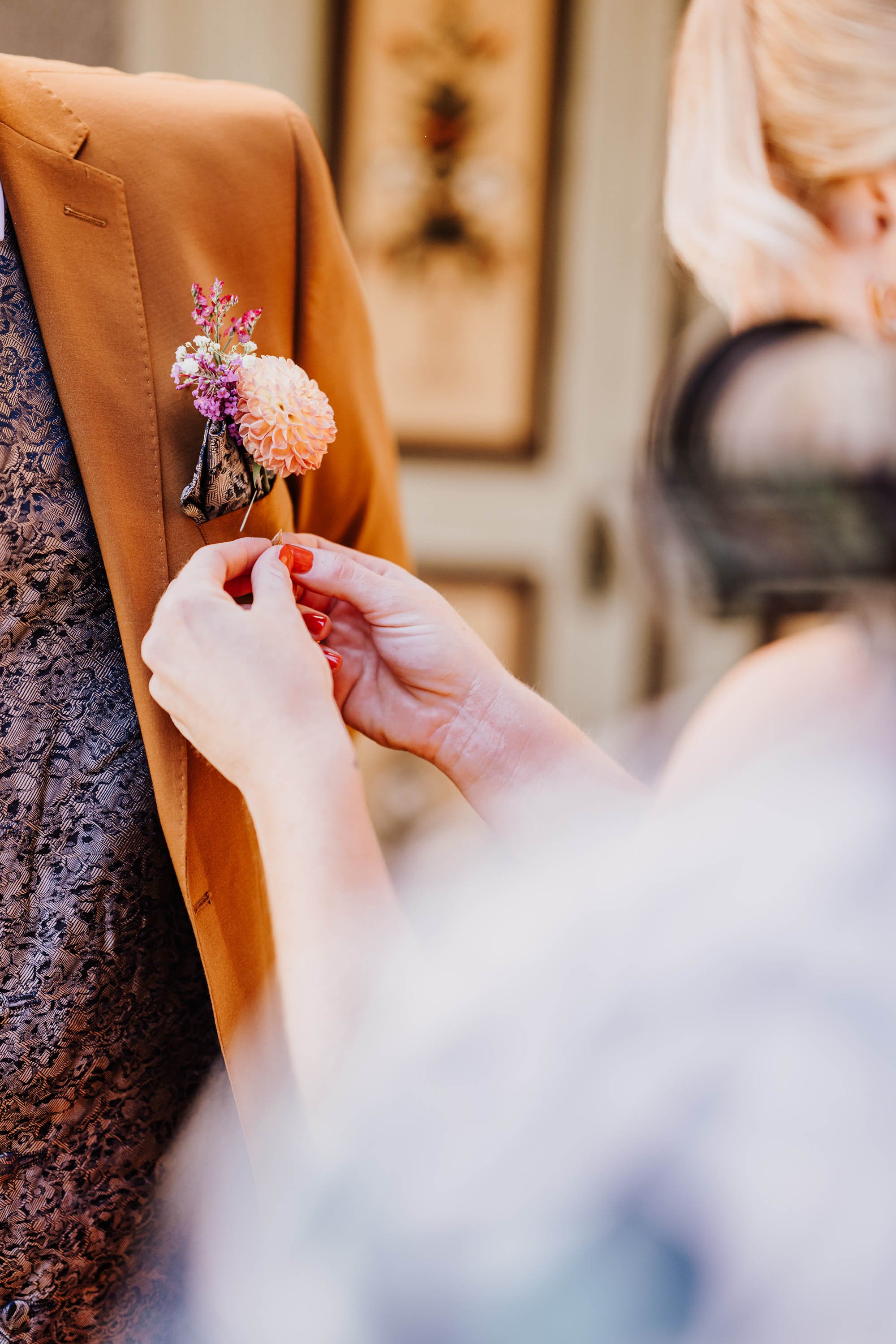 Frau steckt eine Blume in die Brusttasche eines braunen Sakkos, in das eine zweite Blume gesteckt ist.