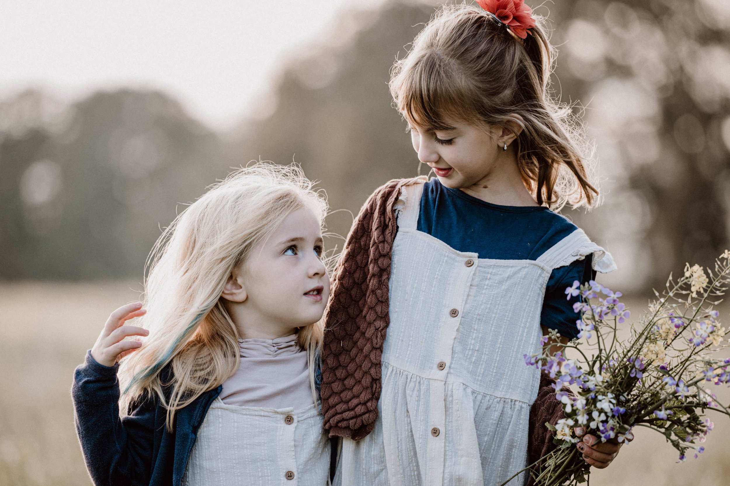 Zwei junge Mädchen in einem Feld, eines hebt die Hand ans Haar, das andere hält einen Blumenstrauß, beide schauen sich liebevoll an.