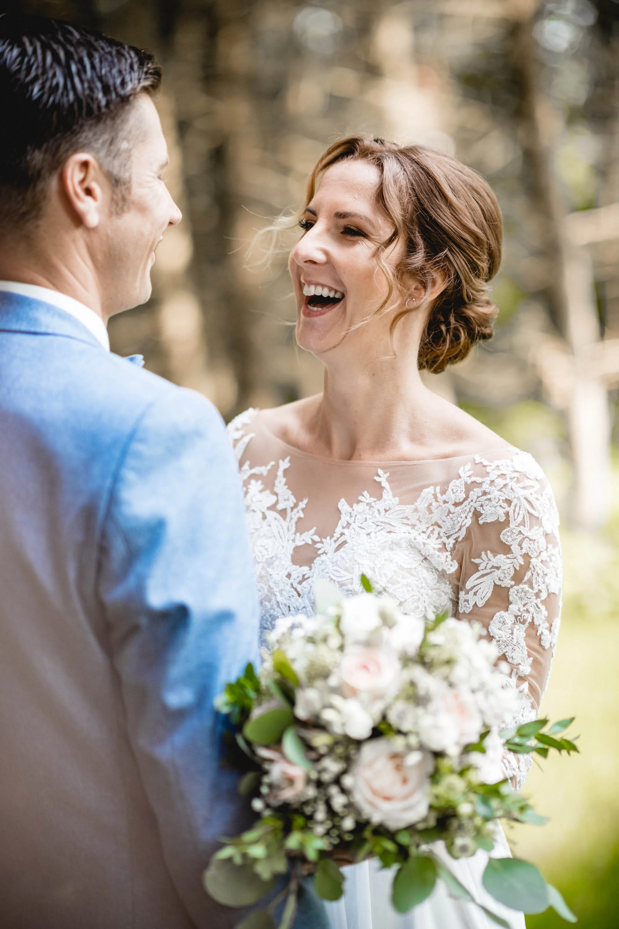 Ein glückliches Brautpaar, das bei Sonnenlicht in einem Wald im Freien lächelt. Die Braut trägt ein weißes Hochzeitskleid mit Spitzendetails und hält einen Blumenstrauß, während der Bräutigam in einem hellblauen Anzug steht.
