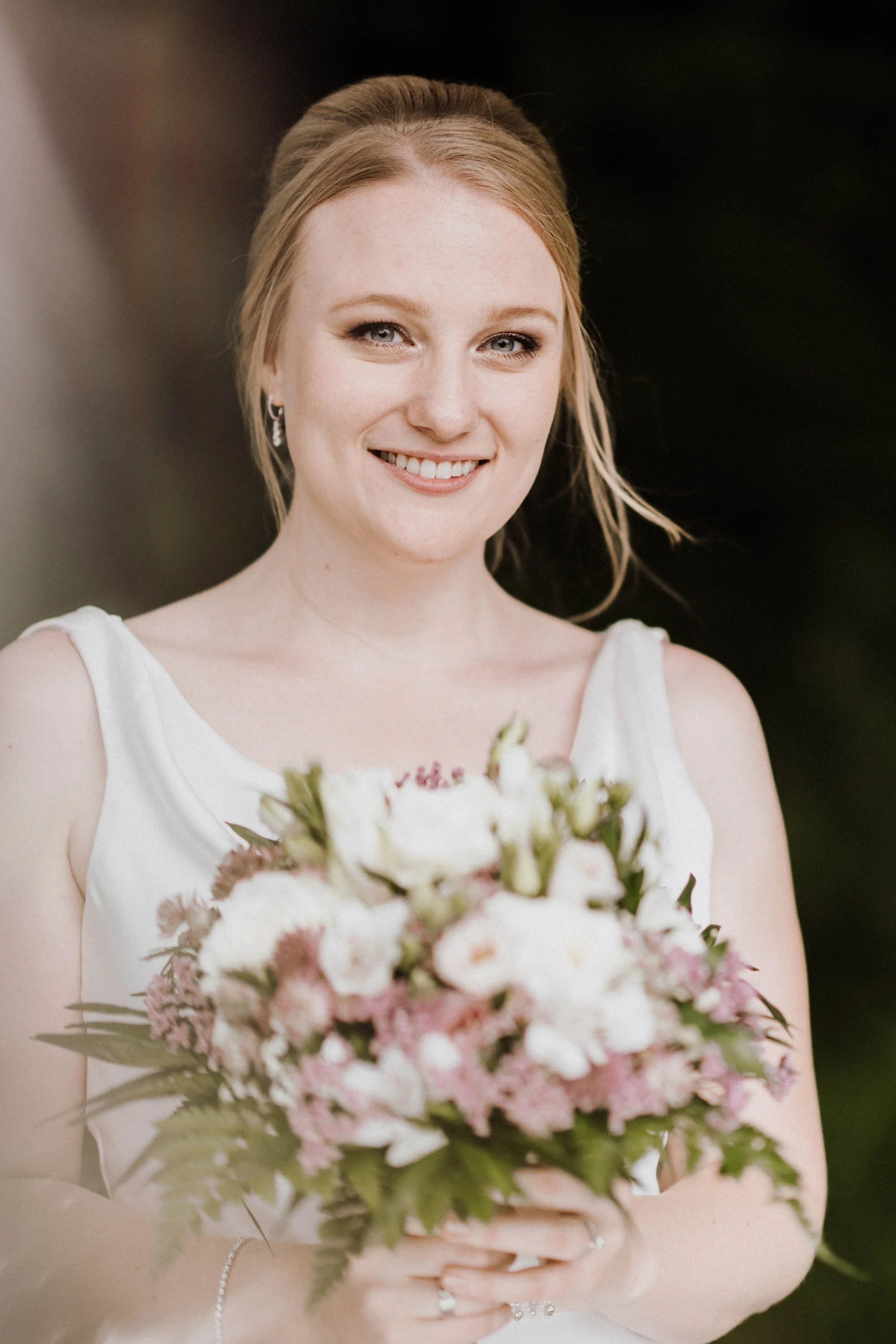 Eine lächelnde Frau in einem weißen Kleid hält einen Blumenstrauß bei einer Hochzeit, im Hintergrund ist ein dunkler, unscharfer Hintergrund zu sehen.