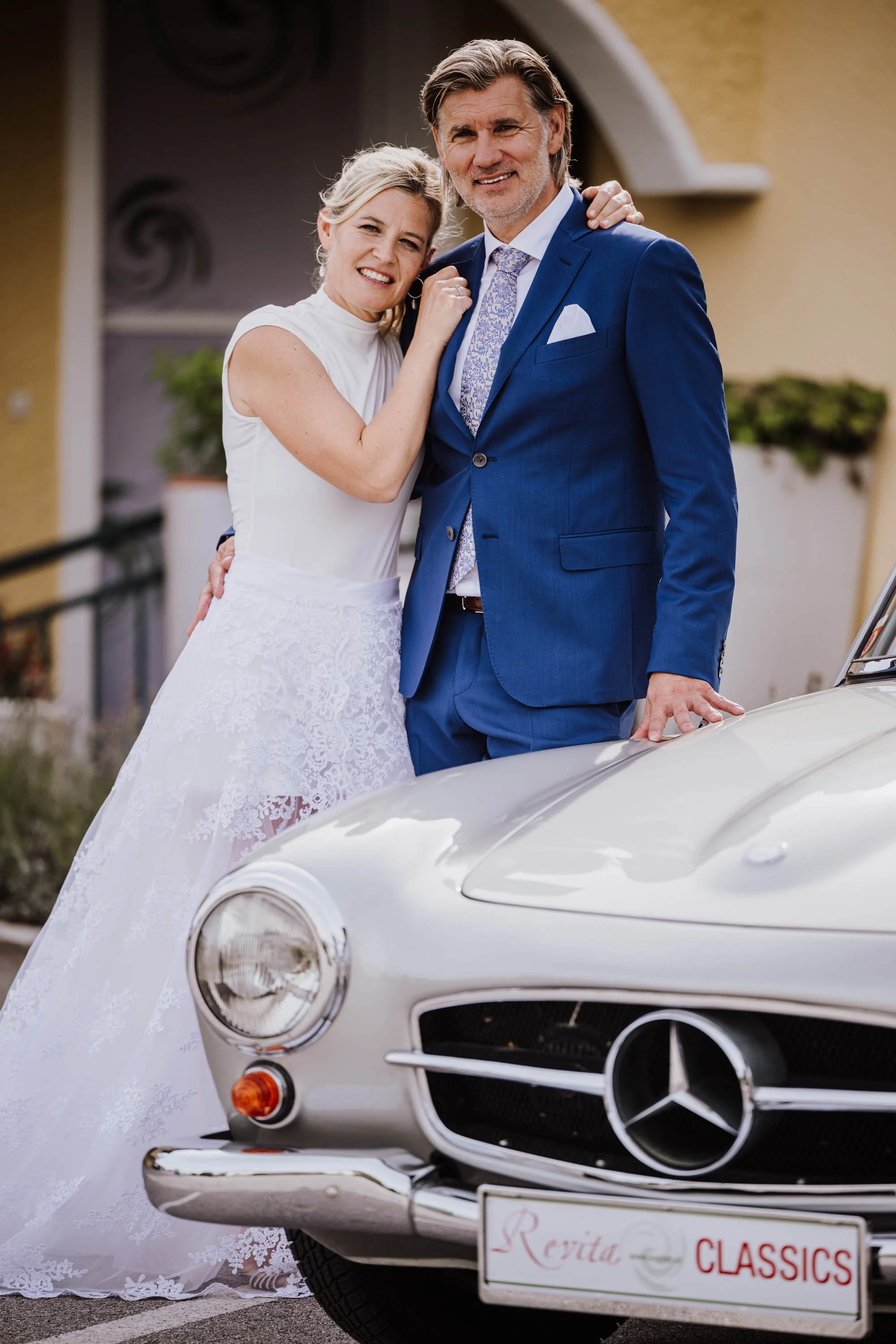 Ein Hochzeitsfoto mit einem älteren Brautpaar in formeller Kleidung, das vor einem klassischen Mercedes-Benz Auto steht, vor einer gelben Hauswand. Die Frau trägt ein weißes Hochzeitskleid und lächelt, während der Mann im blauen Anzug steht.