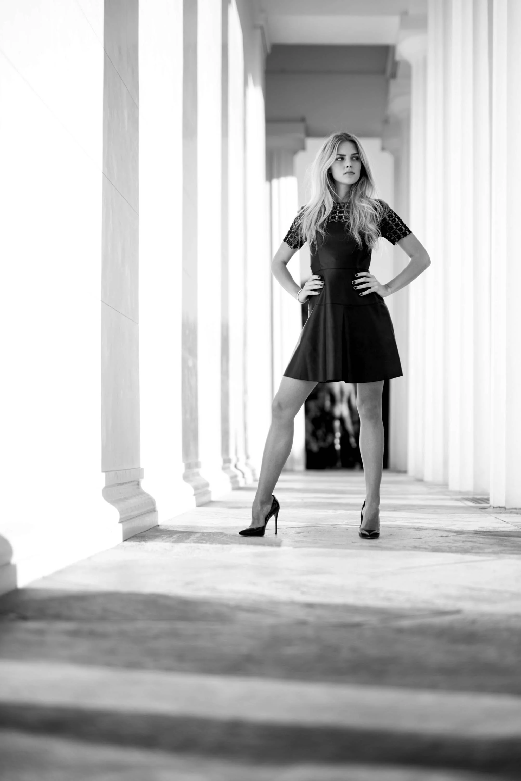 Junge Frau in eleganter schwarzer Kleidung mit High Heels steht in einem Säulengang, Blick nach rechts, Hände in der Hüfte.