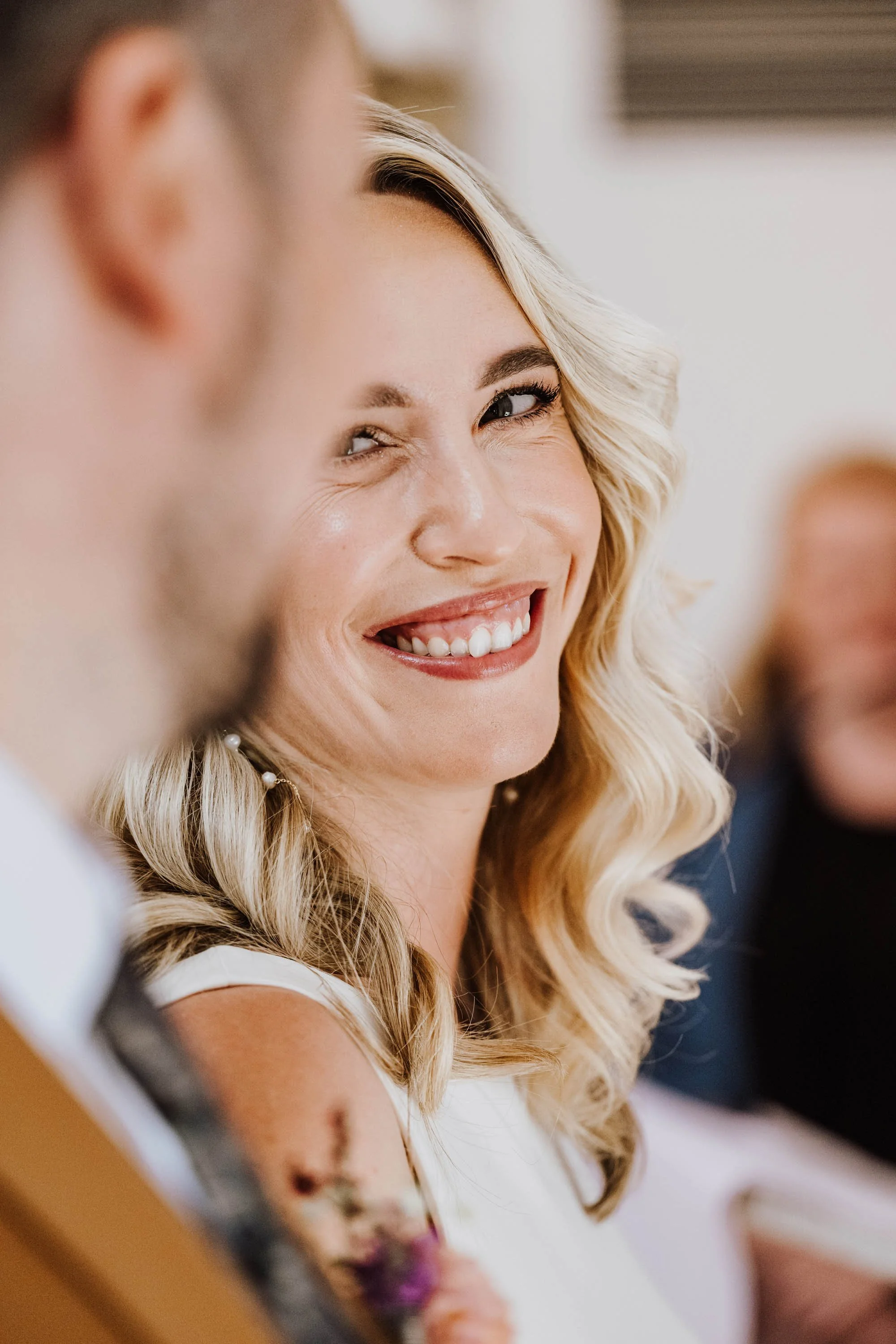 Lächelnde blonde Frau mit lockigem Haar, die auf einer Feier oder in einer freundlichen Gesellschaft sitzt.