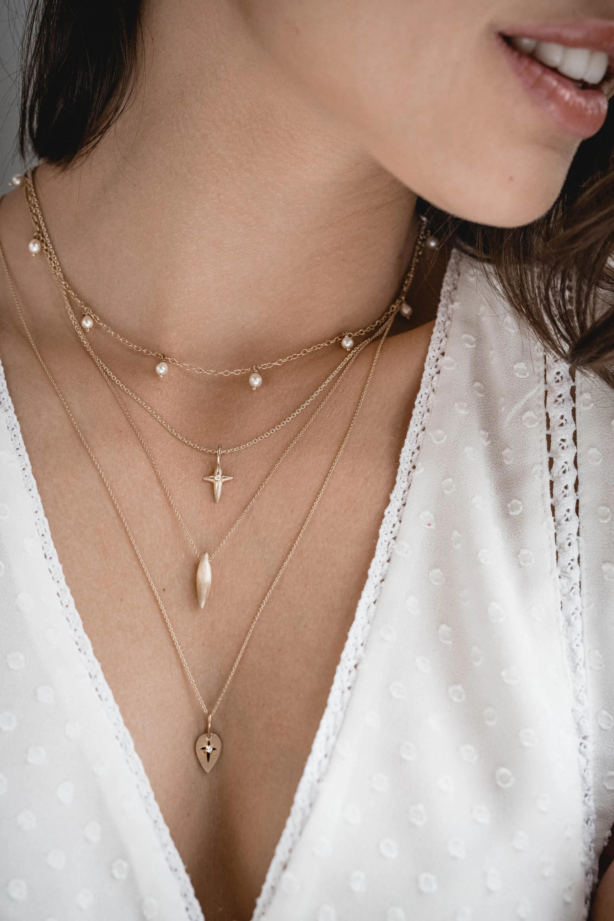 Nahaufnahme von mehreren goldenen Halsketten mit Perlchen und Anhängern, getragen über einem weißen, ausgestanzten Top.