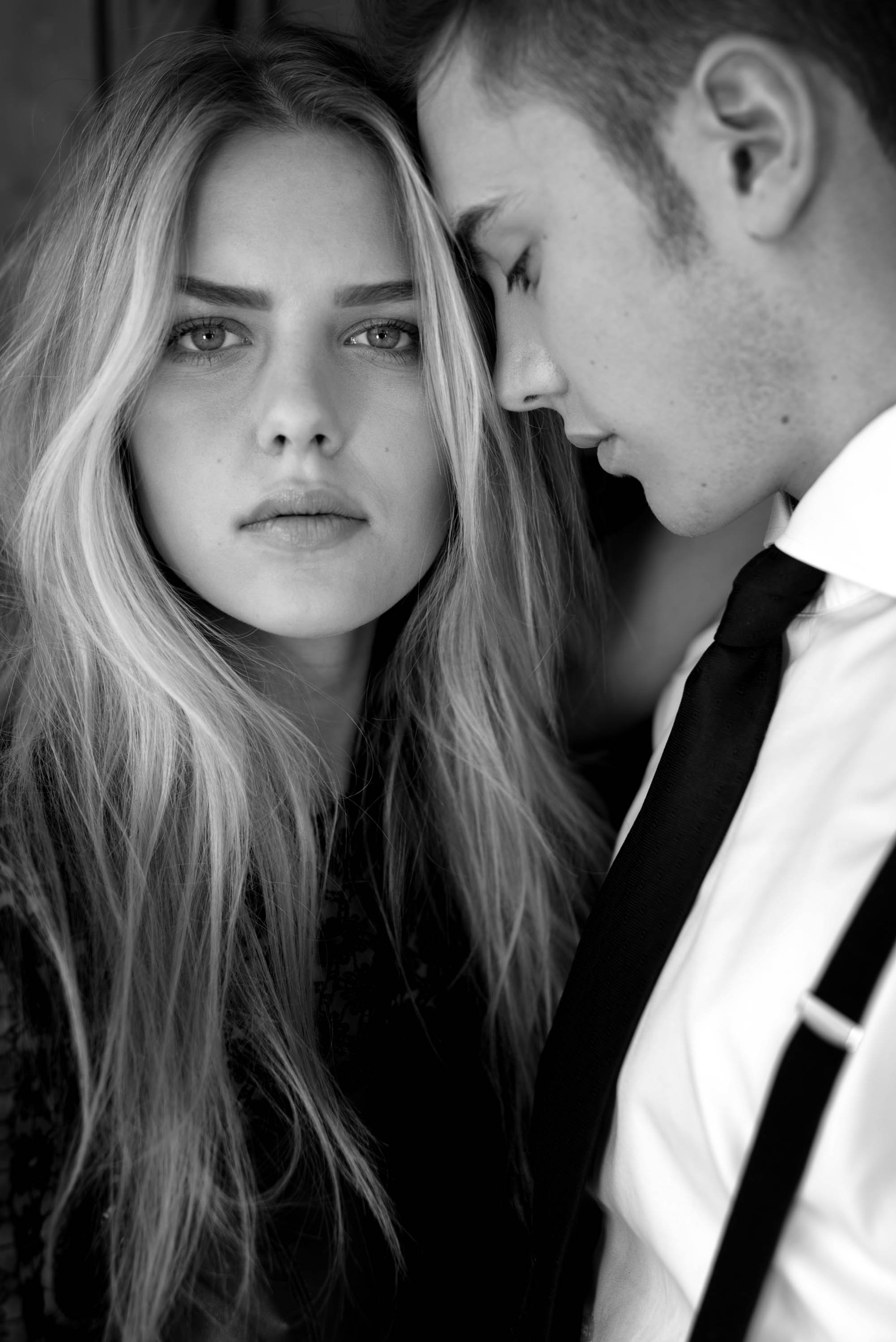 Ein Schwarz-Weiß-Foto eines Paares, das sich sehr nahe steht. Die Frau blickt direkt in die Kamera, während der Mann sein Gesicht an ihr anschmiegt.