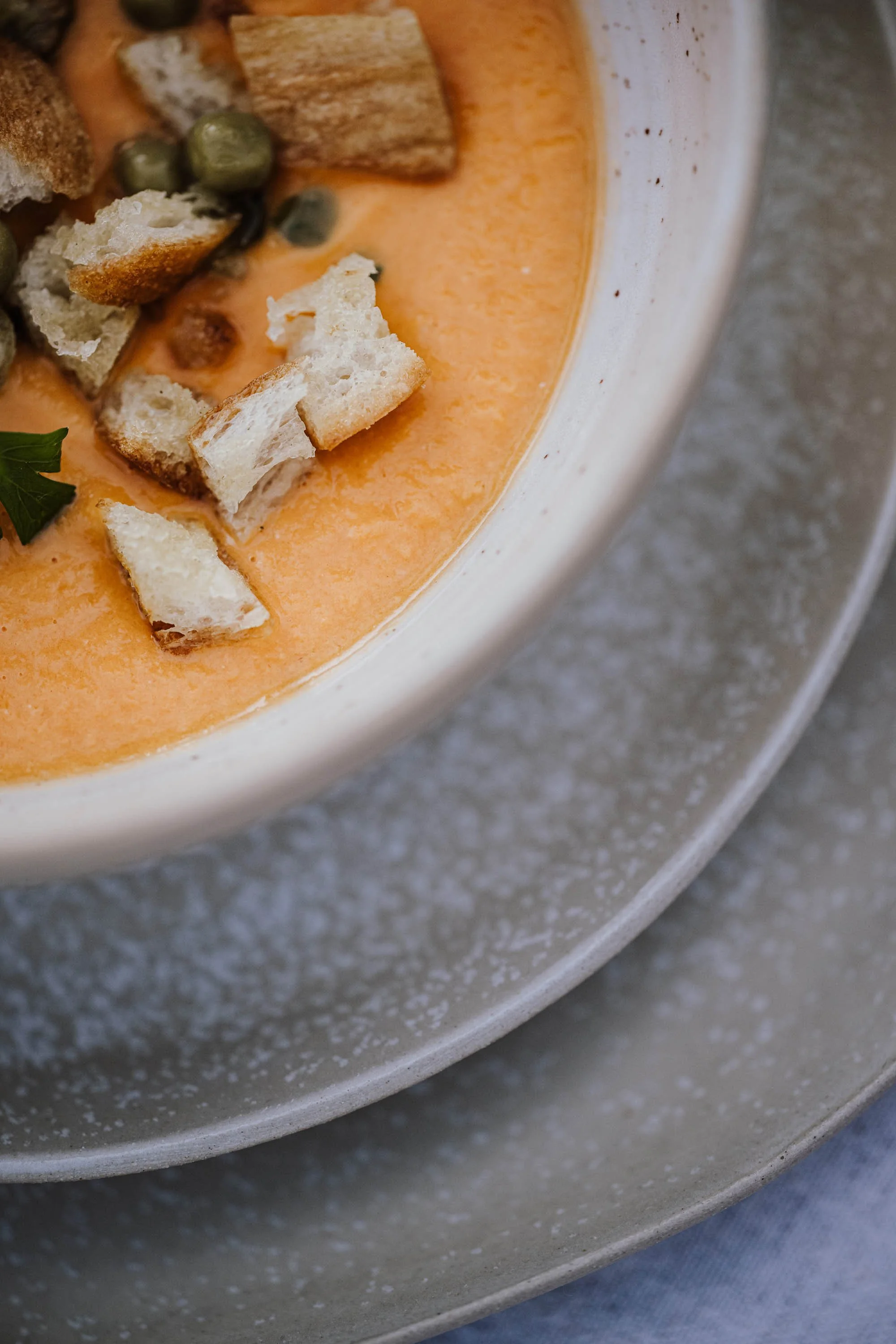 In einer weißen Schüssel befindet sich eine cremige, orangefarbene Suppe, garniert mit Croutons, grünen Kapern und einer grünen Petersilienblatt.