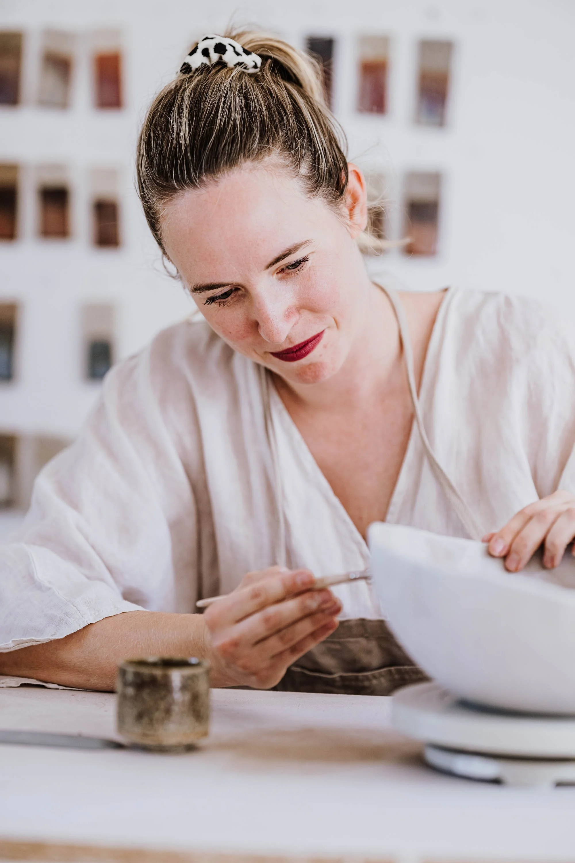 Eine Frau mit roten Lippen und einem weißem Hemd arbeitet mit einer Pinsel auf einer weißen Keramikschale in einer Kreativwerkstatt.