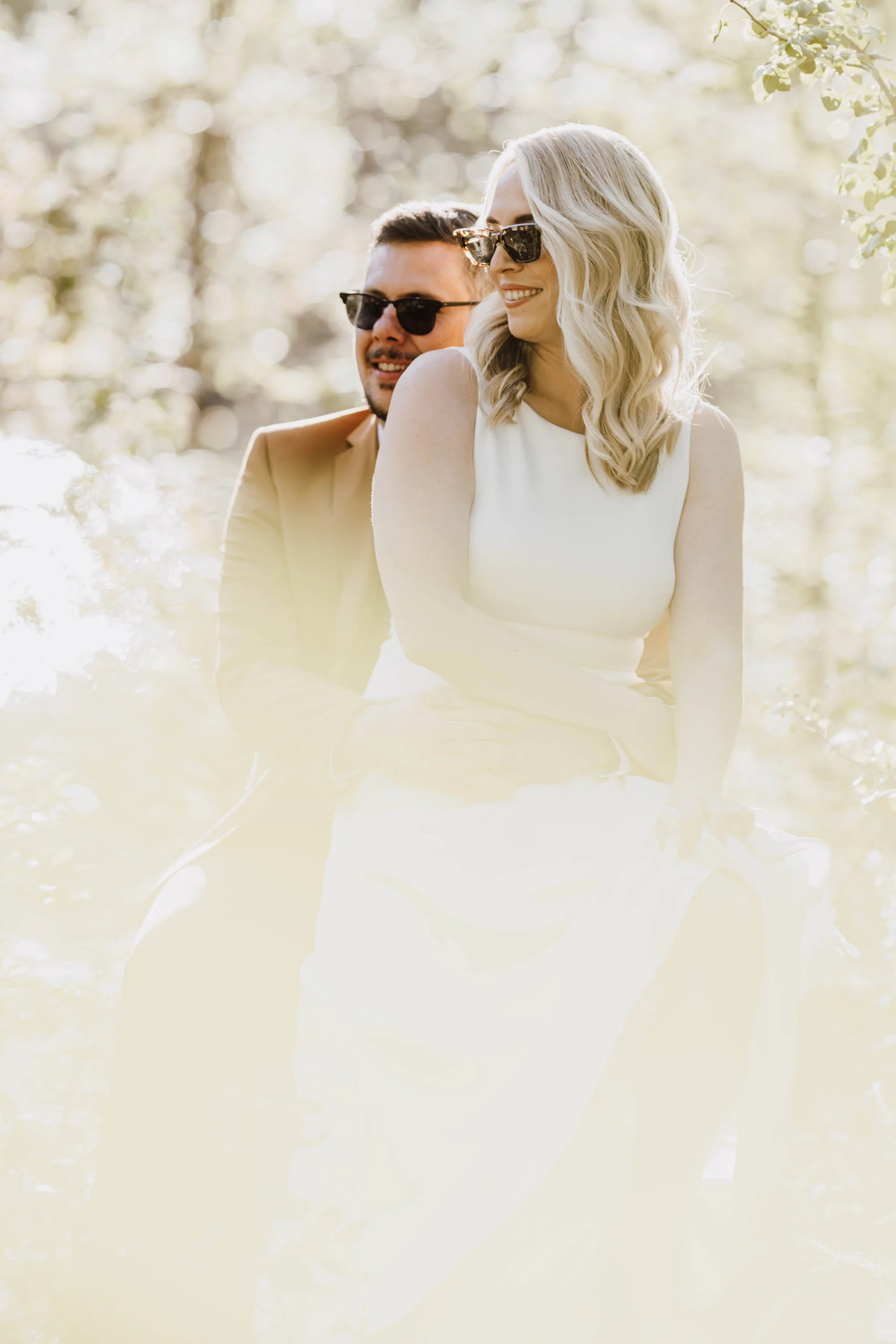 Ein lachendes Paar sitzt eng beieinander im Sonnenlicht im Wald, beide tragen Sonnenbrillen.