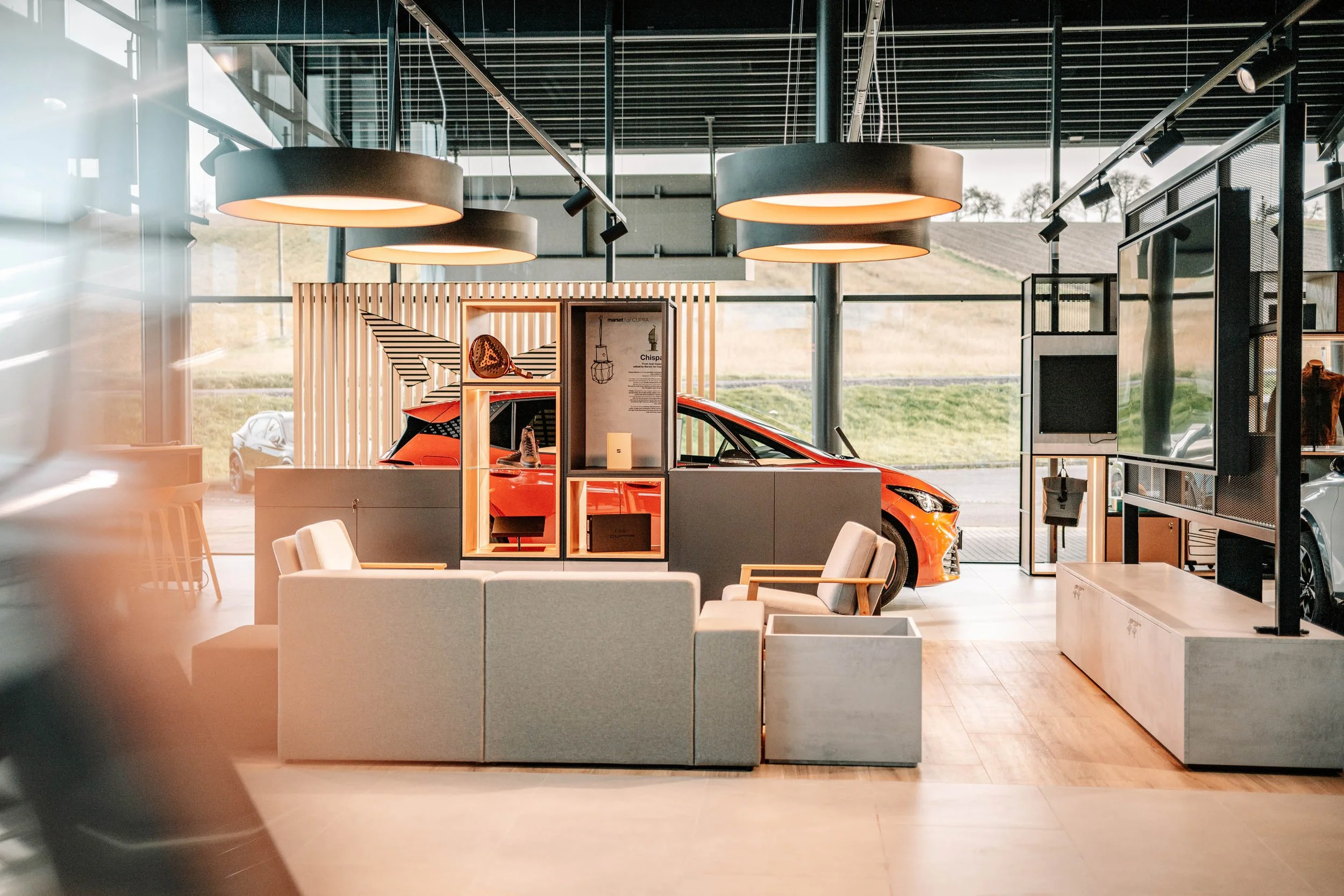 Innenansicht eines Autohauses mit einem roten Auto im Vordergrund, moderner Einrichtung, großer Fensterfront, Aussicht auf grüne Hügellandschaft im Hintergrund.
