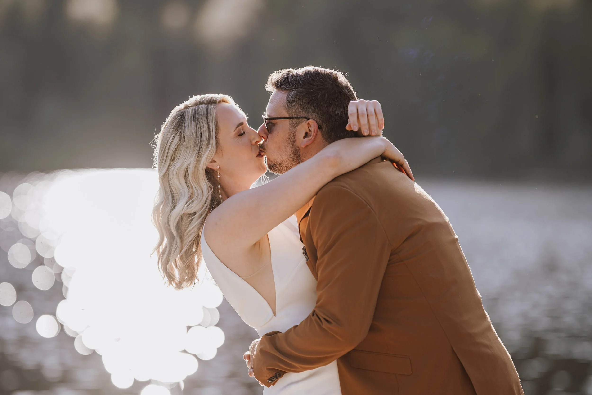 Ein Paar küsst sich am Wasser bei Sonnenuntergang, der Mann trägt eine braune Jacke, die Frau trägt ein weißes Kleid.