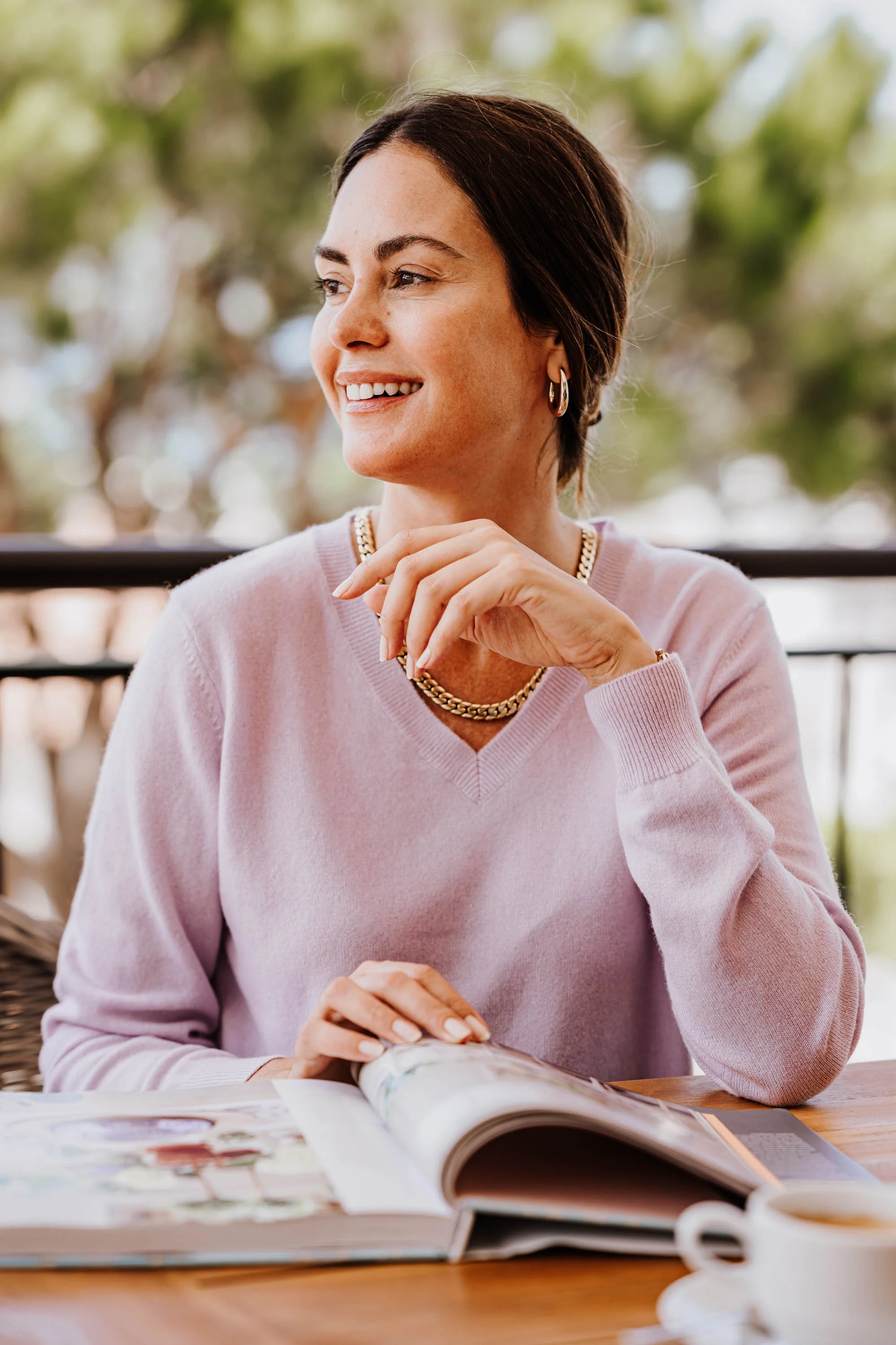 Eine Frau mit kurzen braunen Haaren, die eine rosa Pullover trägt, sitzt an einem Holztisch mit einem offenen Magazin, einem weißen Kaffeetasse und einem Löffel, wirkt lächelnd und entspannt, während sie im Freien sitzt, umgeben von unscharfem grünen Hintergrund.