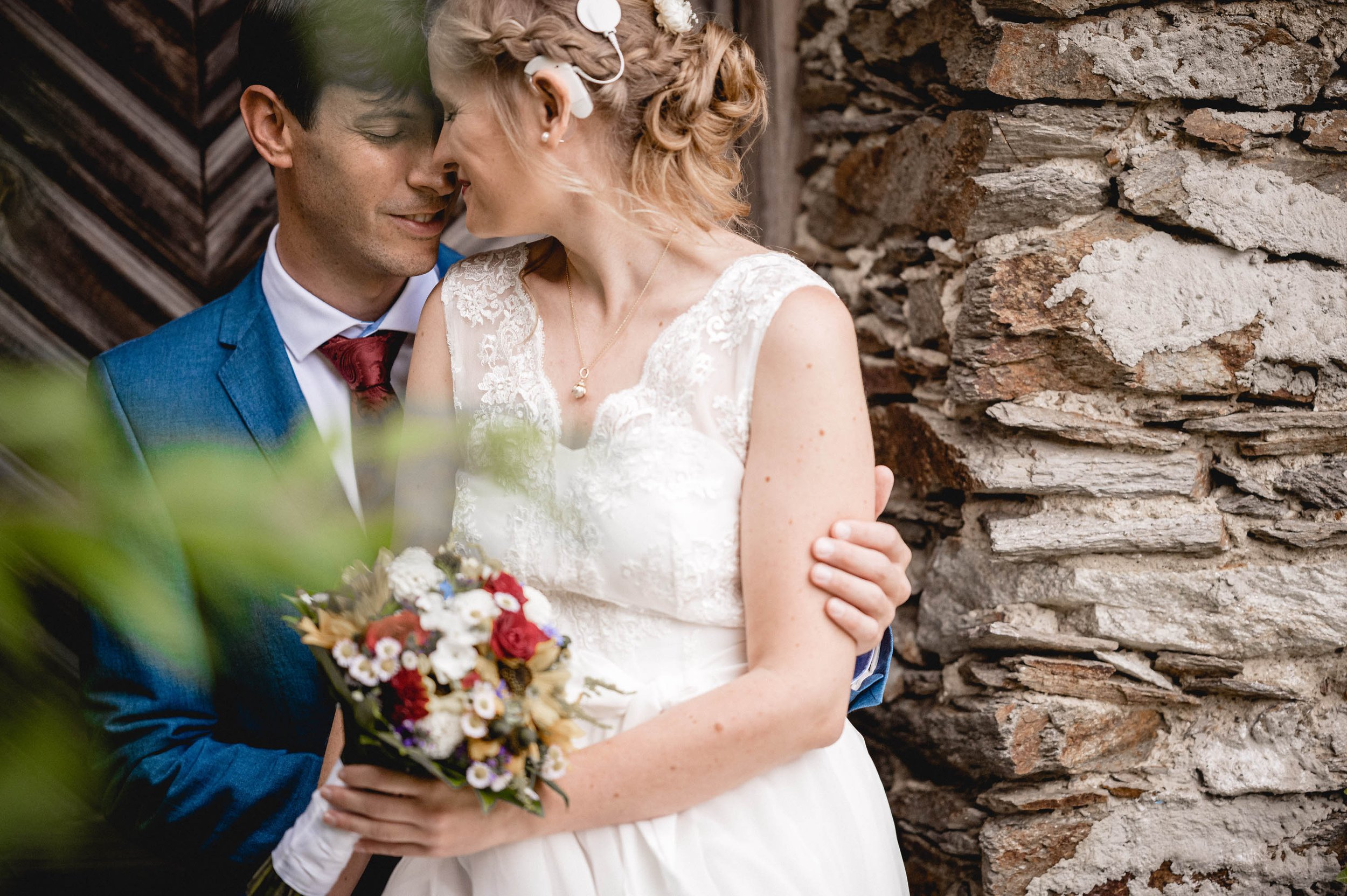 Ein Brautpaar bei einer Hochzeit, umarmend und sich gegenseitig berührend, vor einer alten Steinmauer, die Braut hält einen Blumenstrauß, beide lächeln und sind in festlicher Kleidung gekleidet.