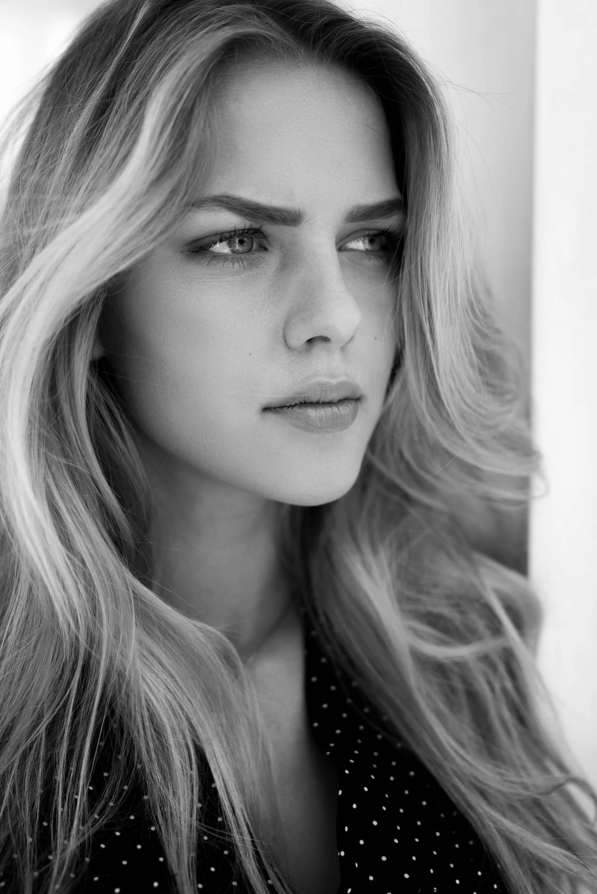 Schwarz-weiß Porträt einer jungen Frau mit freiem, gewellten Haar, die nachdenklich aus dem Fenster schaut.