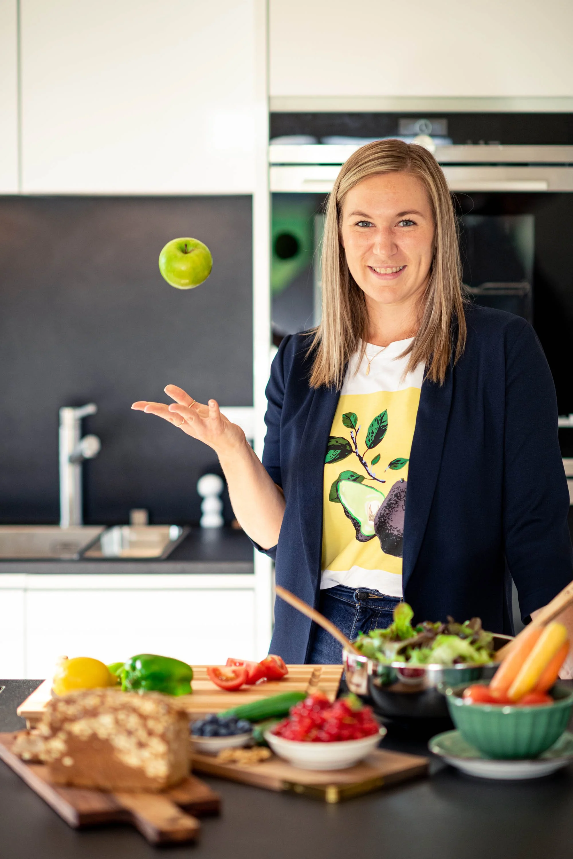 Eine Frau in einer modernen Küche jongliert mit einem grünen Apfel, umgeben von verschiedenen frischen Lebensmitteln auf der Küchenarbeitsplatte.
