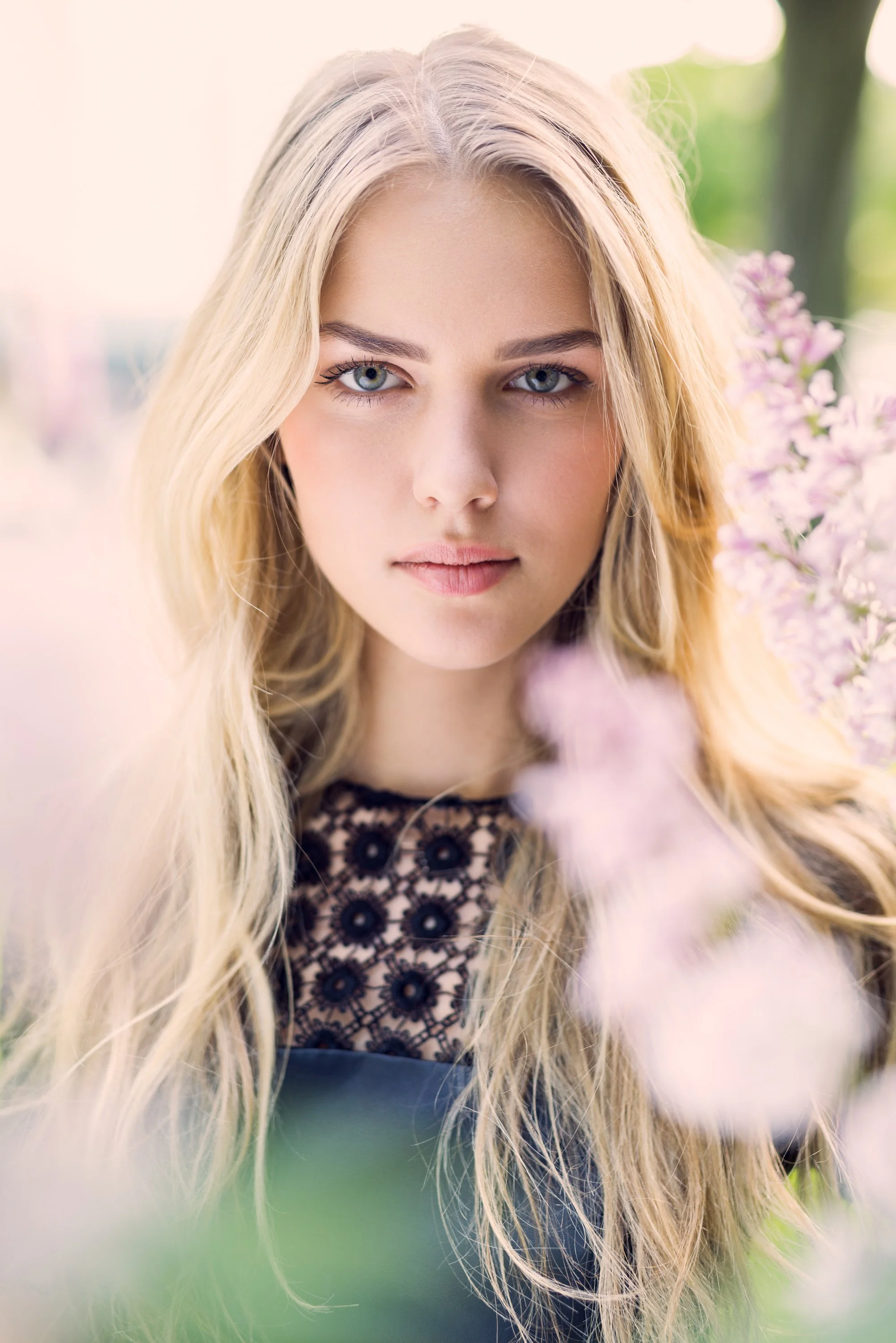 Ein junges Mädchen mit blonden langen Haaren und blauen Augen, posiert vor einer blühenden Pflanze mit rosa Blüten, im Freien bei hellem Tageslicht.