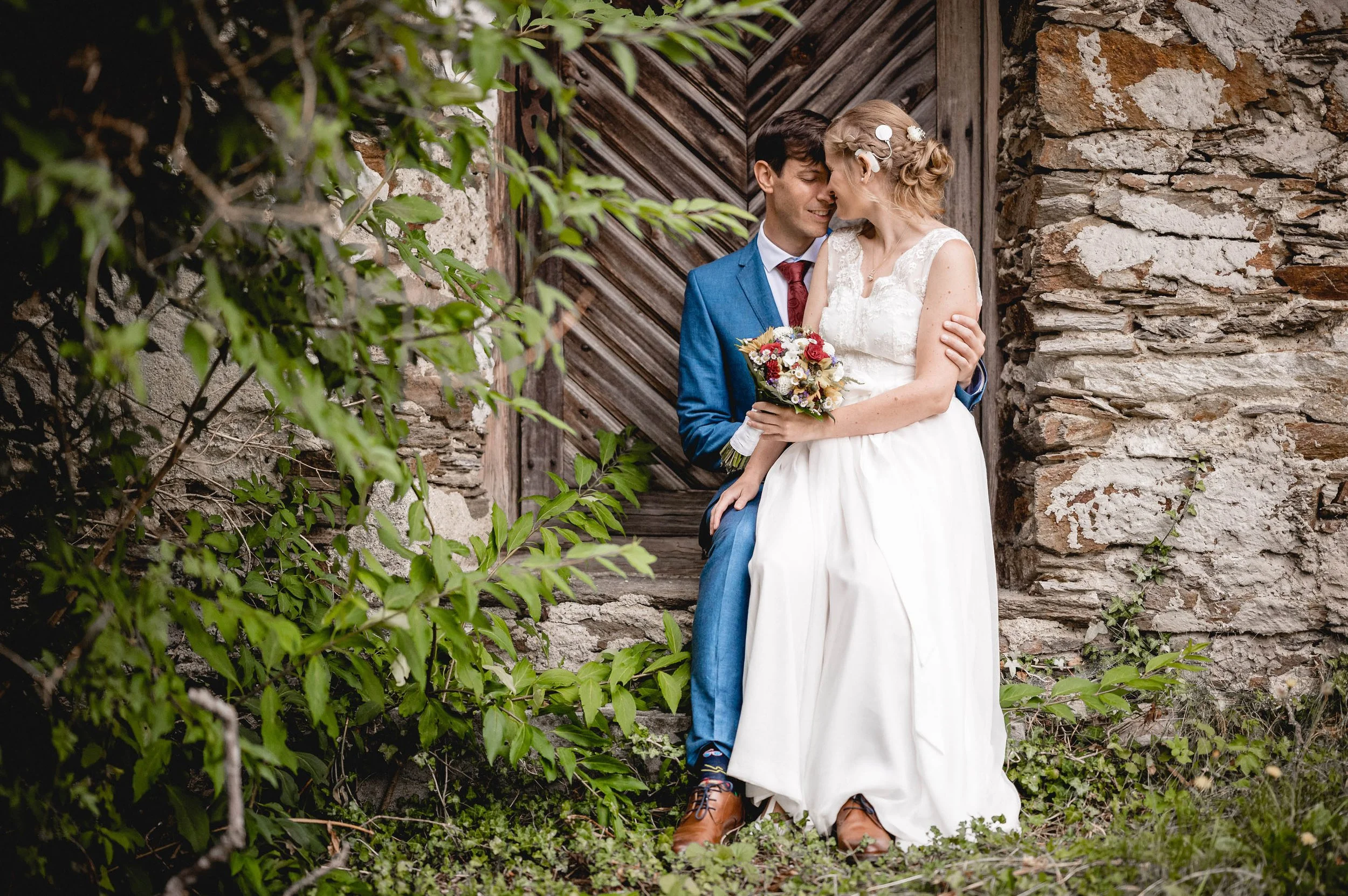 Ein Brautpaar sitzt vor einer alten Steinmauer und einem Holztor, umarmt sich, die Braut hält einen Blumenstrauß, beide lächeln sich an.