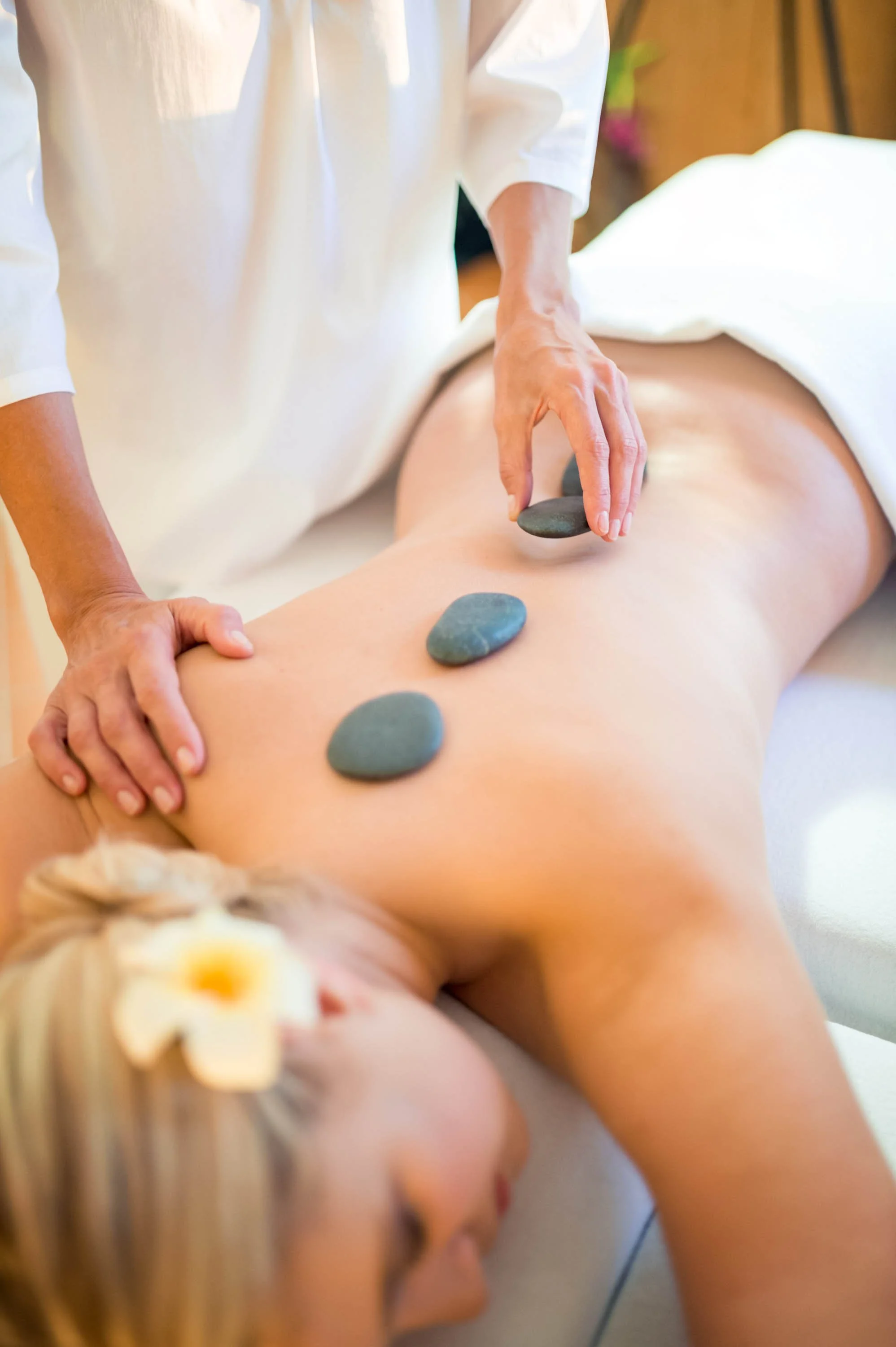 Eine Person erhält eine Hot-Stone-Massage auf ihrem Rücken in einem Spa. Steine liegen auf dem Rücken und die Masseurin hält einen Stein in der Hand.
