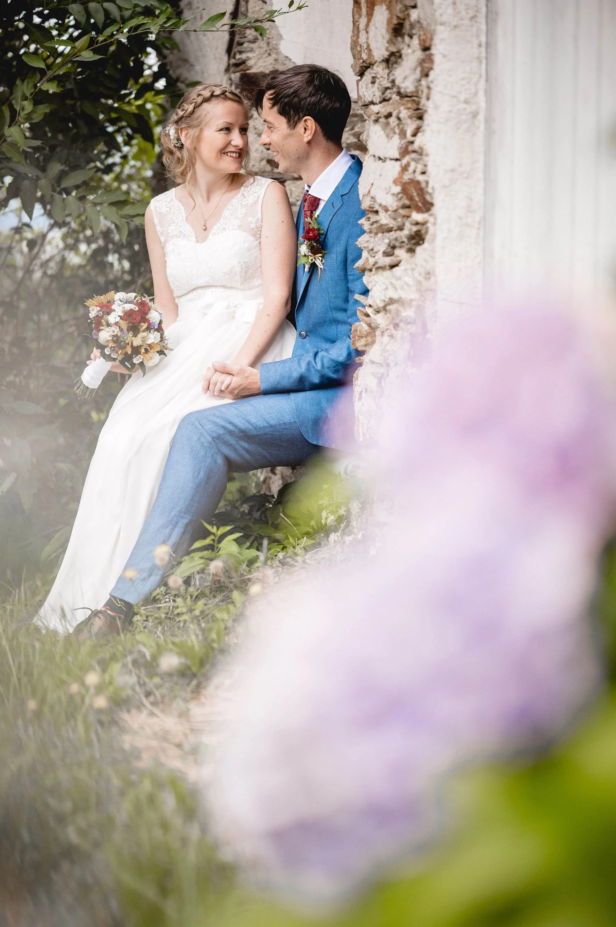 Ein Brautpaar sitzt verliebt im Garten bei einer Hochzeit, umgeben von Blumen und einer alten Steinmauer.