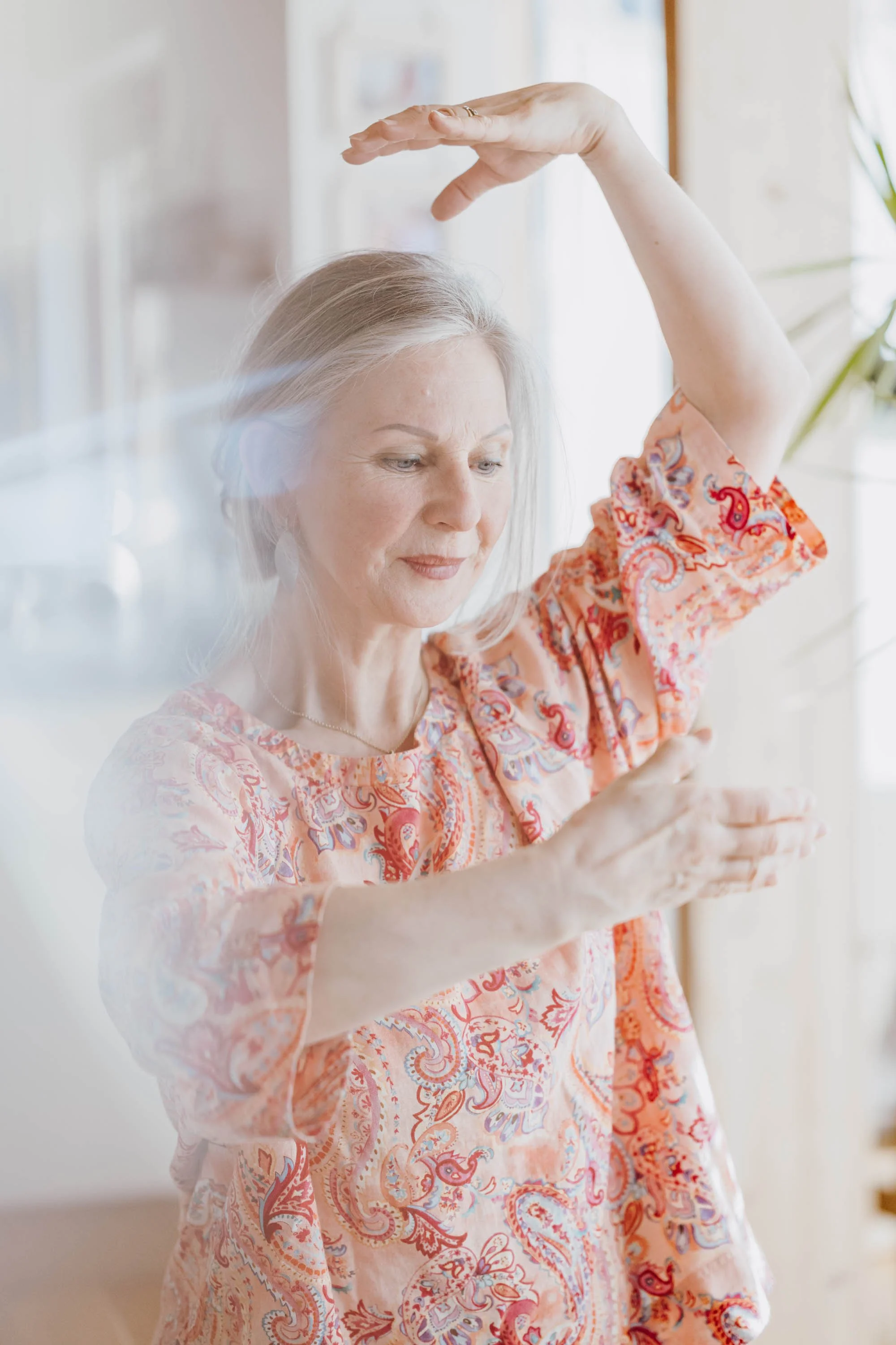 Eine ältere Frau macht eine Tanzbewegung, die Arme und Hände in einer bestimmten Haltung, vor einem hellen, lichtdurchfluteten Hintergrund.