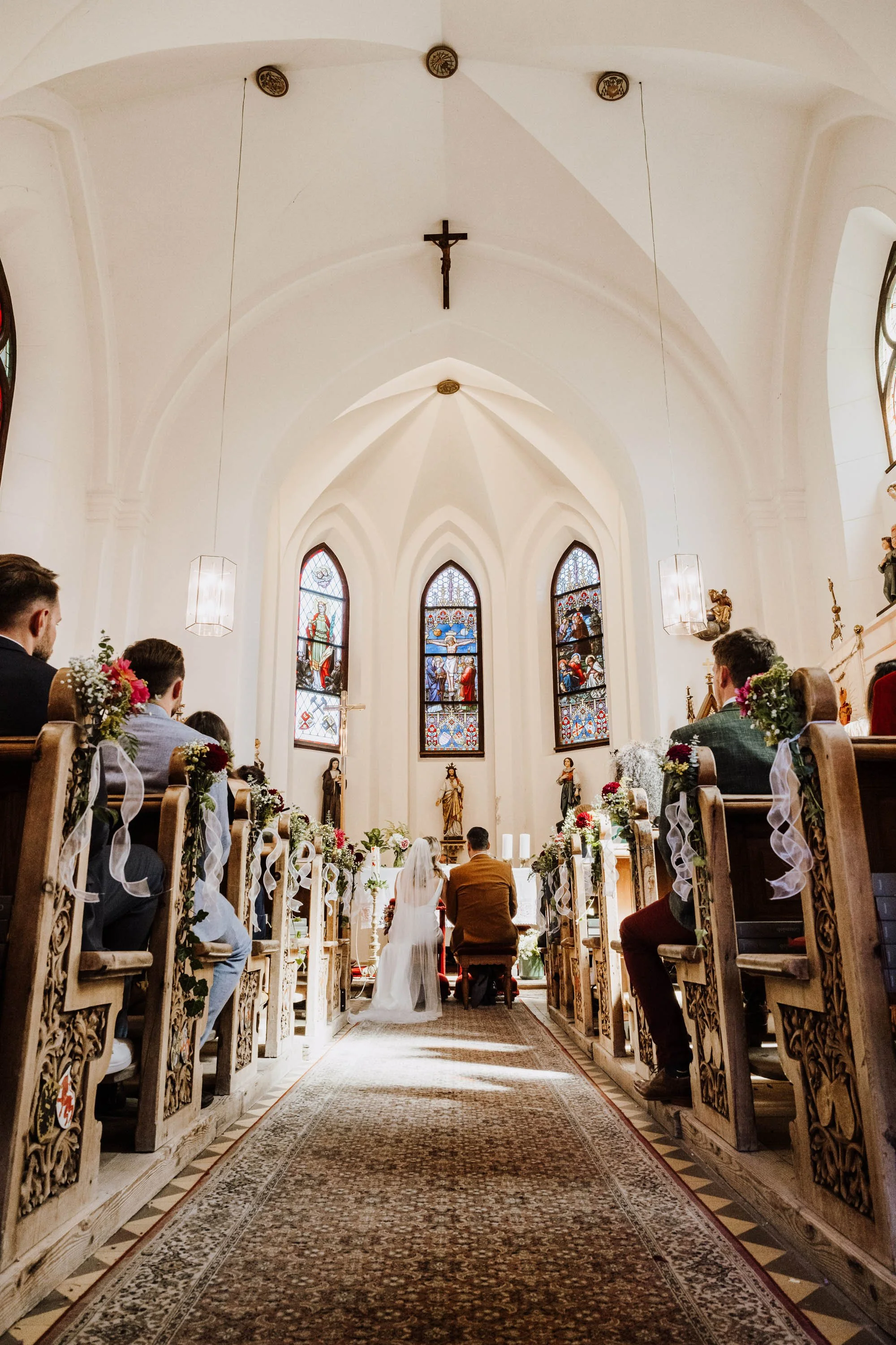 Hochzeit in einer Kirche mit Braut und Bräutigam vor dem Altar, umgeben von Hochzeitsgästen, with Kirchenfenstern und religiösen Statuen im Hintergrund.