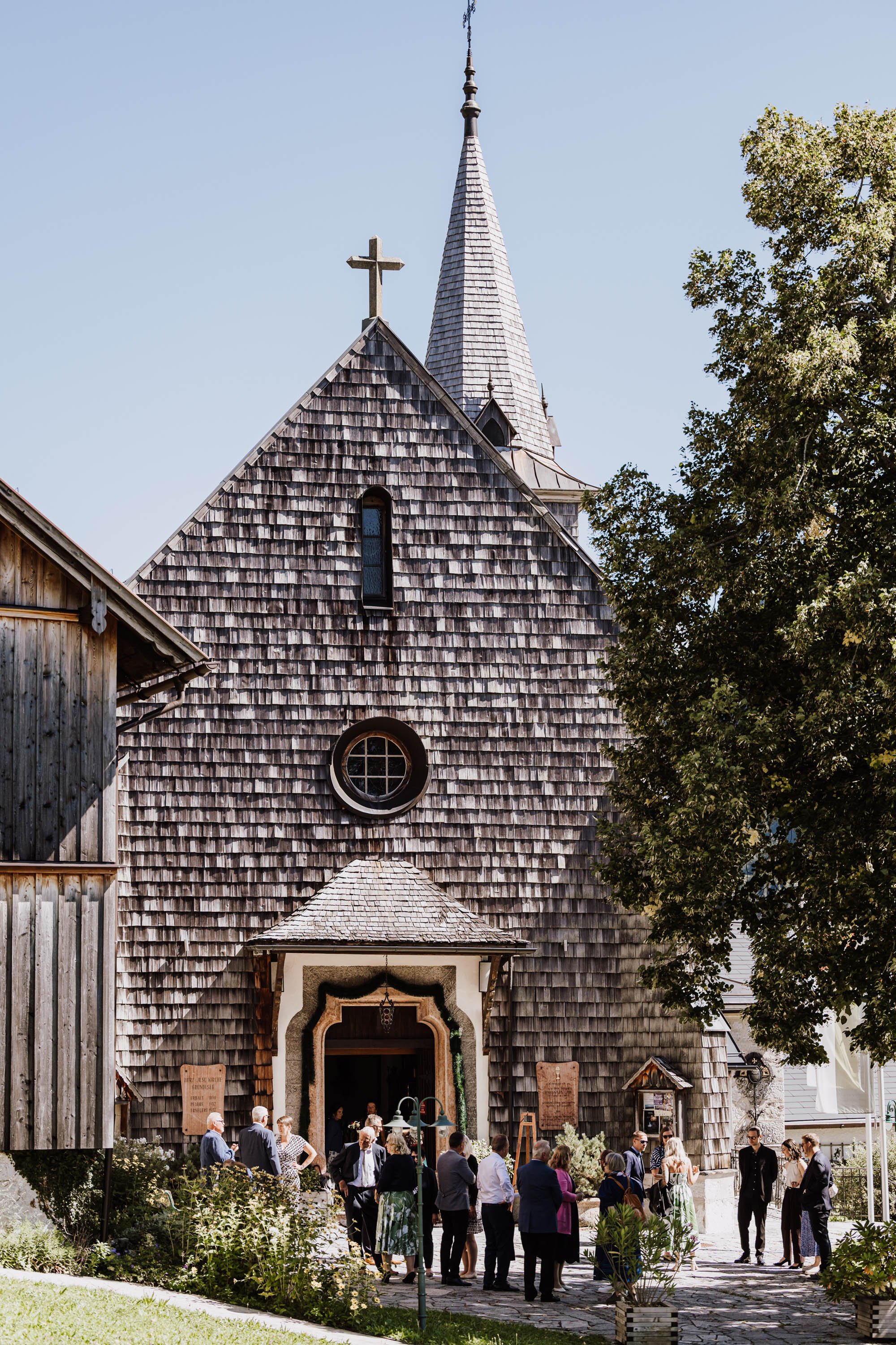 Eine Holzkirche mit steilem Dach und einem Turm mit Spitze, umgeben von Bäumen, mit Leuten vor der Tür, die sich versammeln.