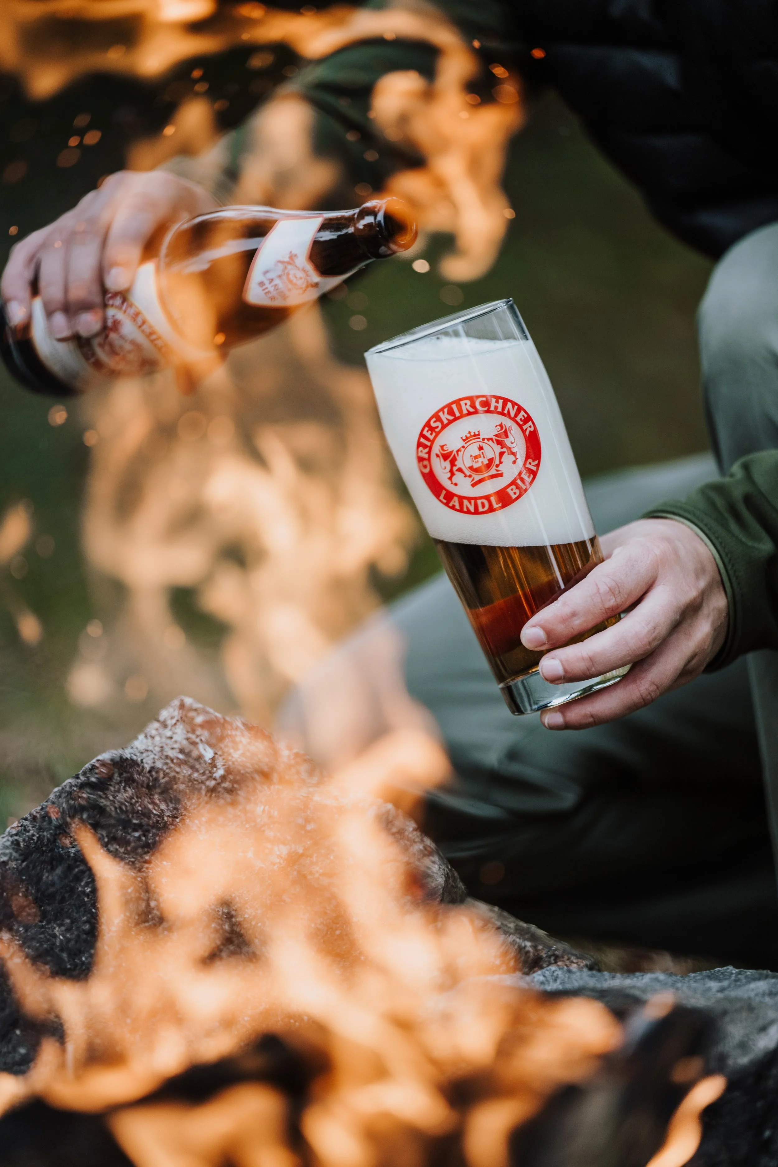 Person, die Bier in einem Glas mit dem Logo 'GareisKirchner Landl Bier' einschenkt, während ein Lagerfeuer im Vordergrund brennt.