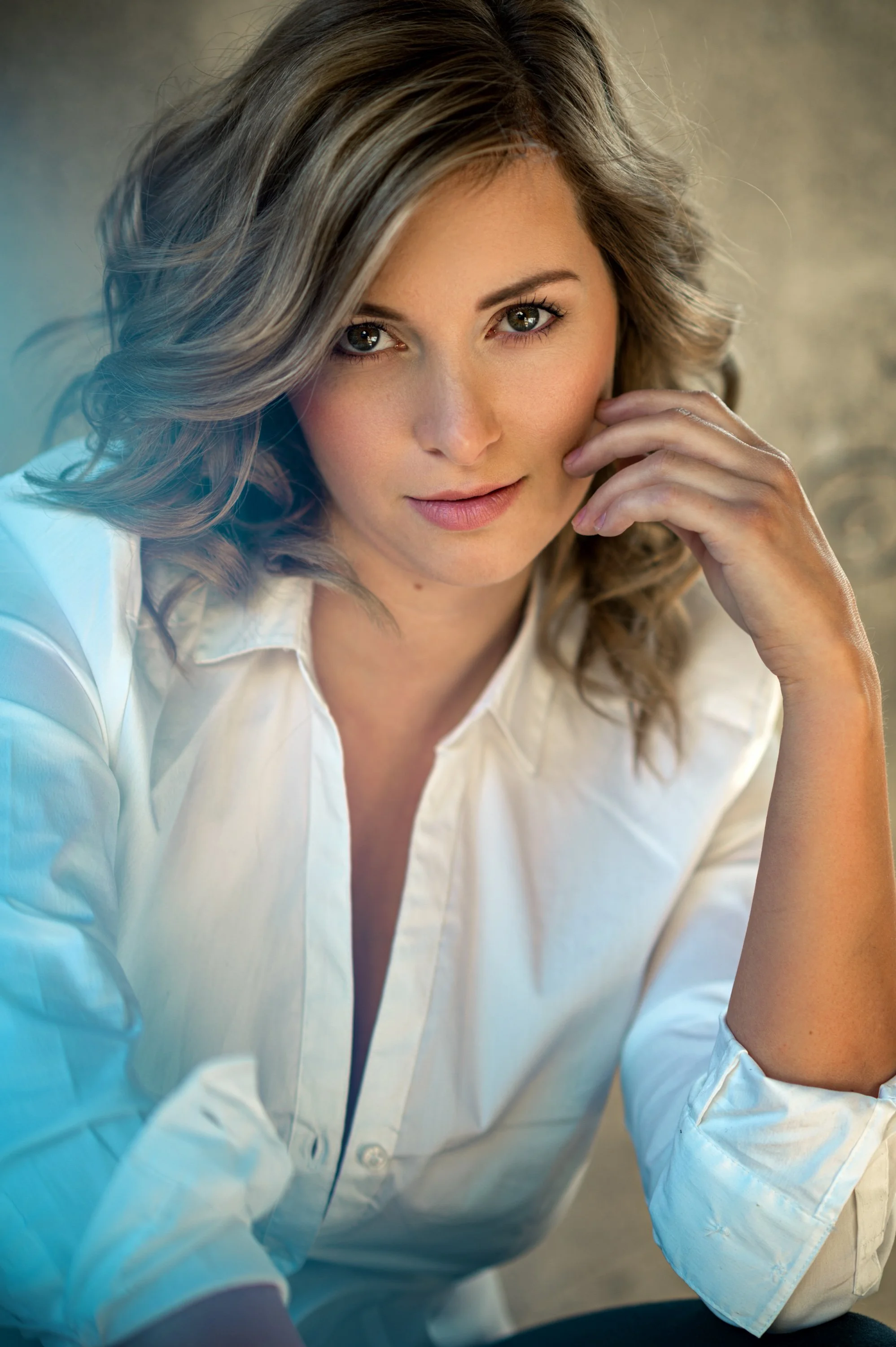 Nahaufnahme einer jungen Frau mit lockigem, blondem Haar, die ein weißes Hemd trägt. Sie schaut direkt in die Kamera, wirkt entspannt und freundlich, legt ihre Hand an ihr Gesicht.