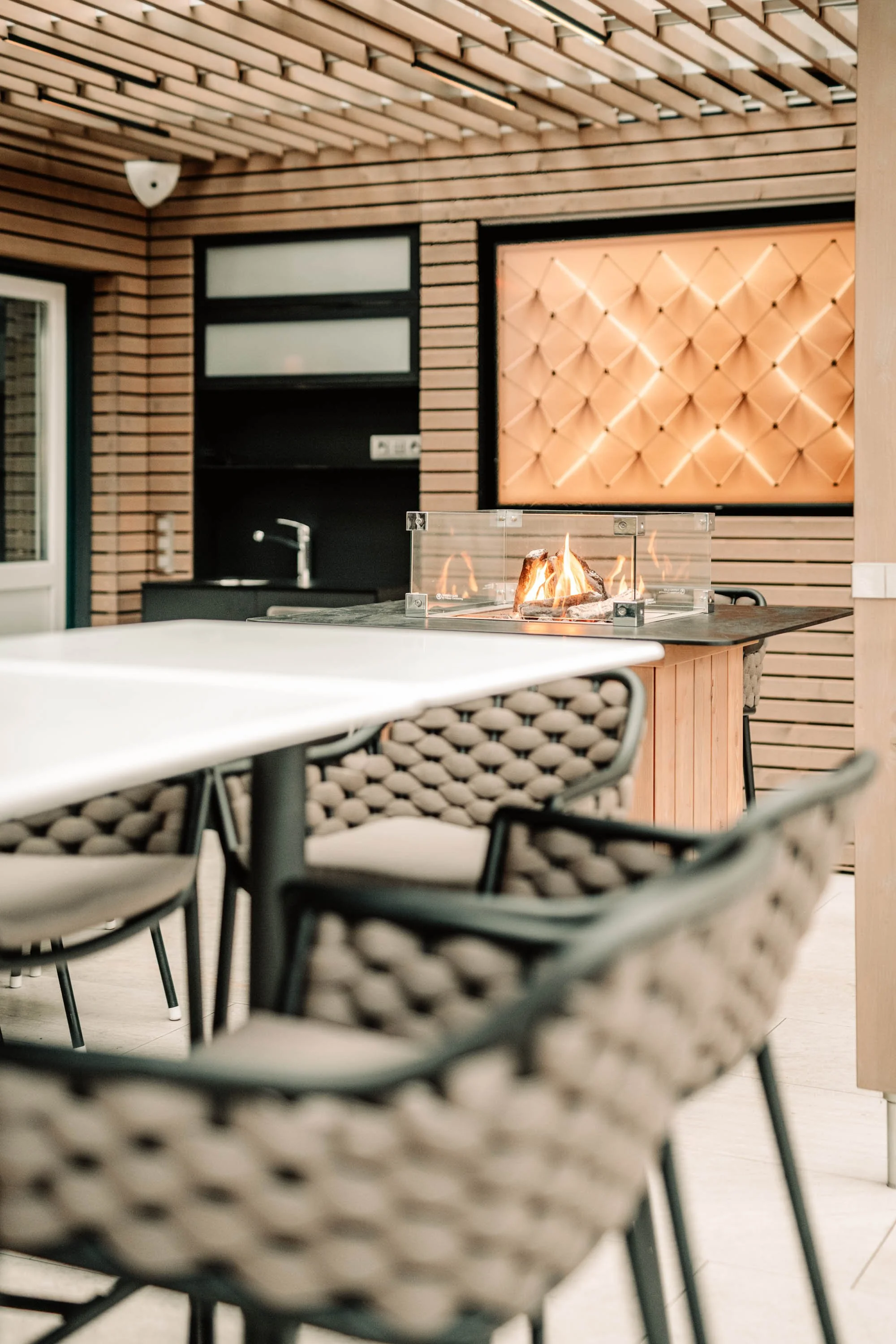 Ein moderner Innenhof mit einem Holzdeck, einem Tisch mit einem entflammbaren Feuer auf einer Glasbox, umgeben von Stühlen mit geflochtenen Sitzflächen.