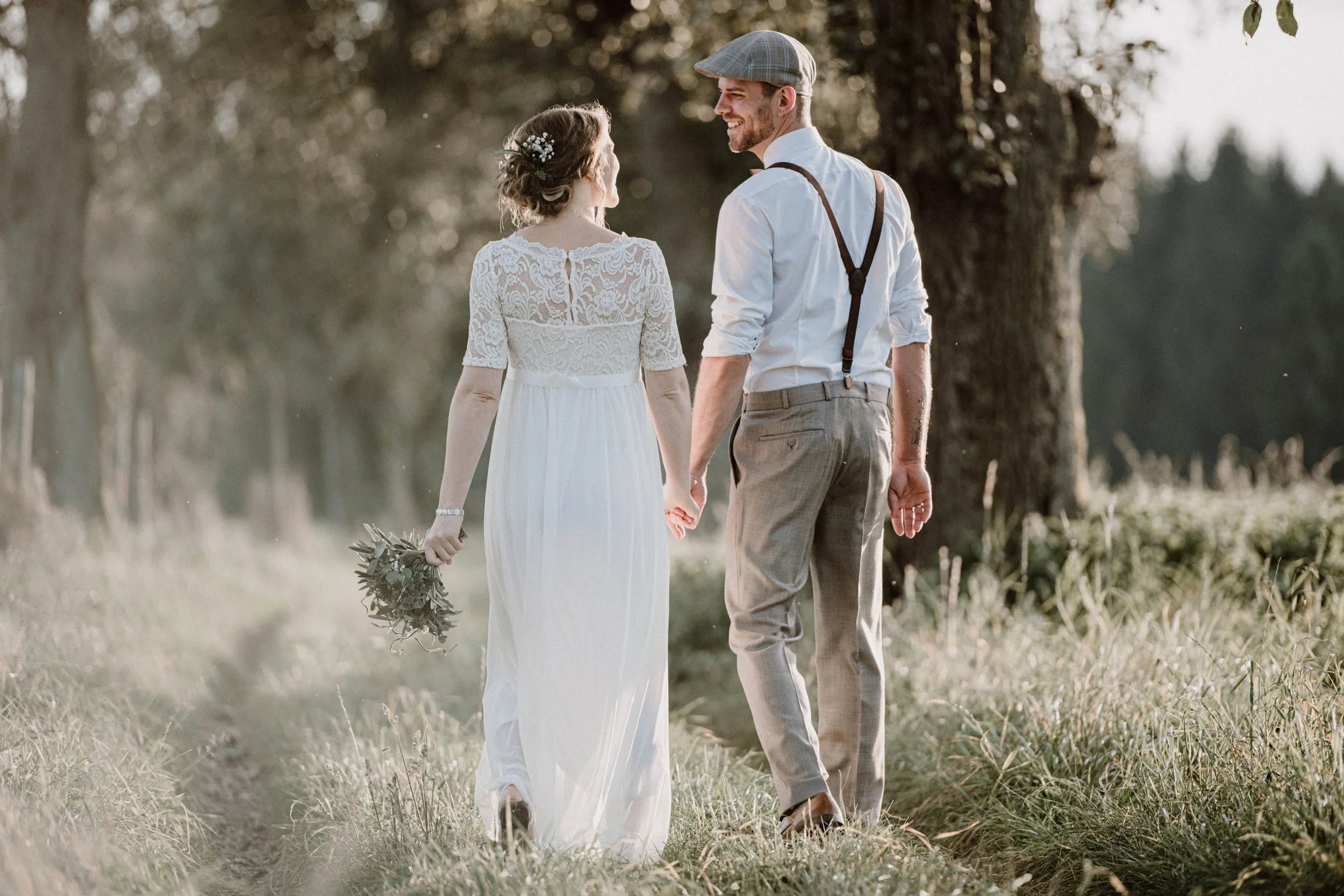 Ein Brautpaar geht Hand in Hand durch eine Wiese bei Sonnenuntergang. Die Frau trägt ein weißes Hochzeitskleid und hält einen Blumenstrauß, der Mann trägt einen weißen Hemd, braune Hose, Hosenträger und einen Kappe.