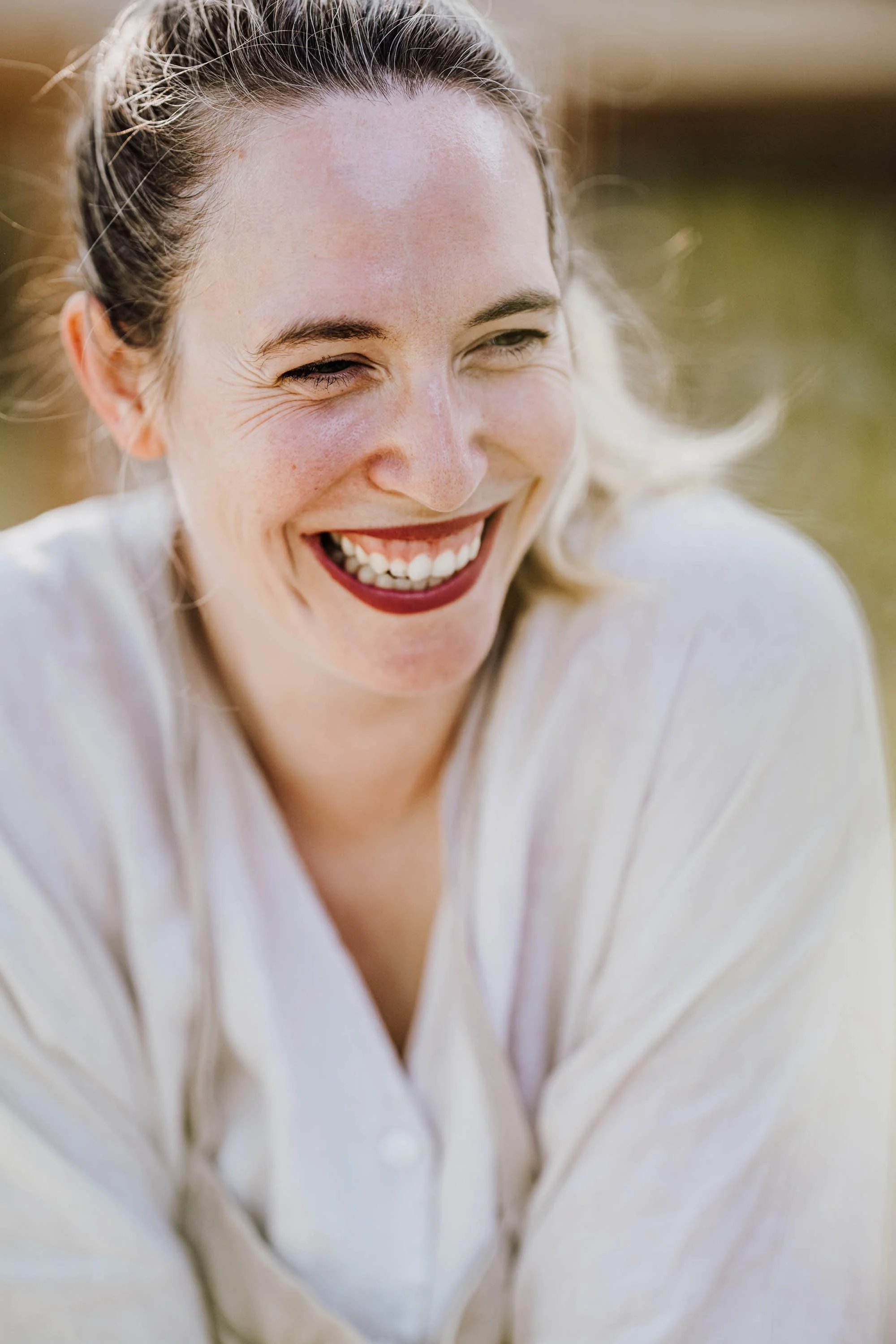 Lächelnde Frau mit offenen Haaren und rotem Lippenstift, sonniger Hintergrund.